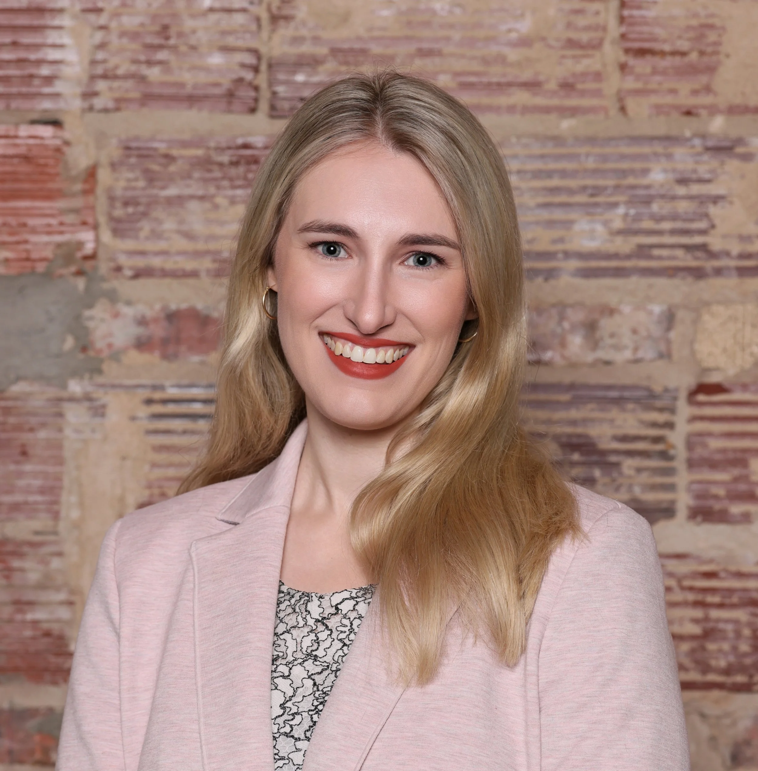 Close-up of a smiling woman with blonde hair and blue eyes, wearing a light pink blazer, standing in front of a brick wall.