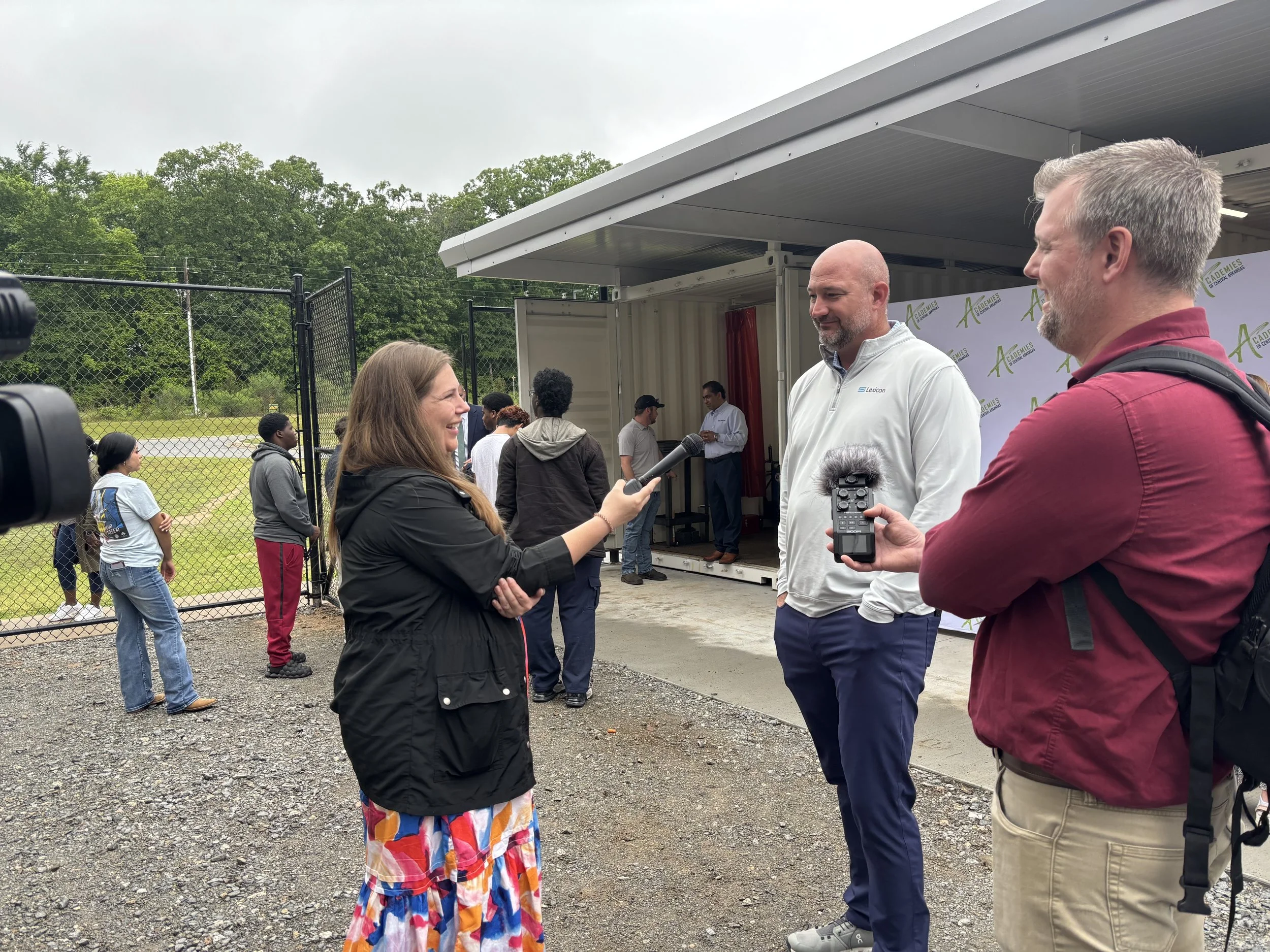 A woman is conducting an interview with a man in an outdoor setting, with a small stage and several people waiting in line in the background. The interviewer holds a microphone, and another man with a backpack is holding a recording device.