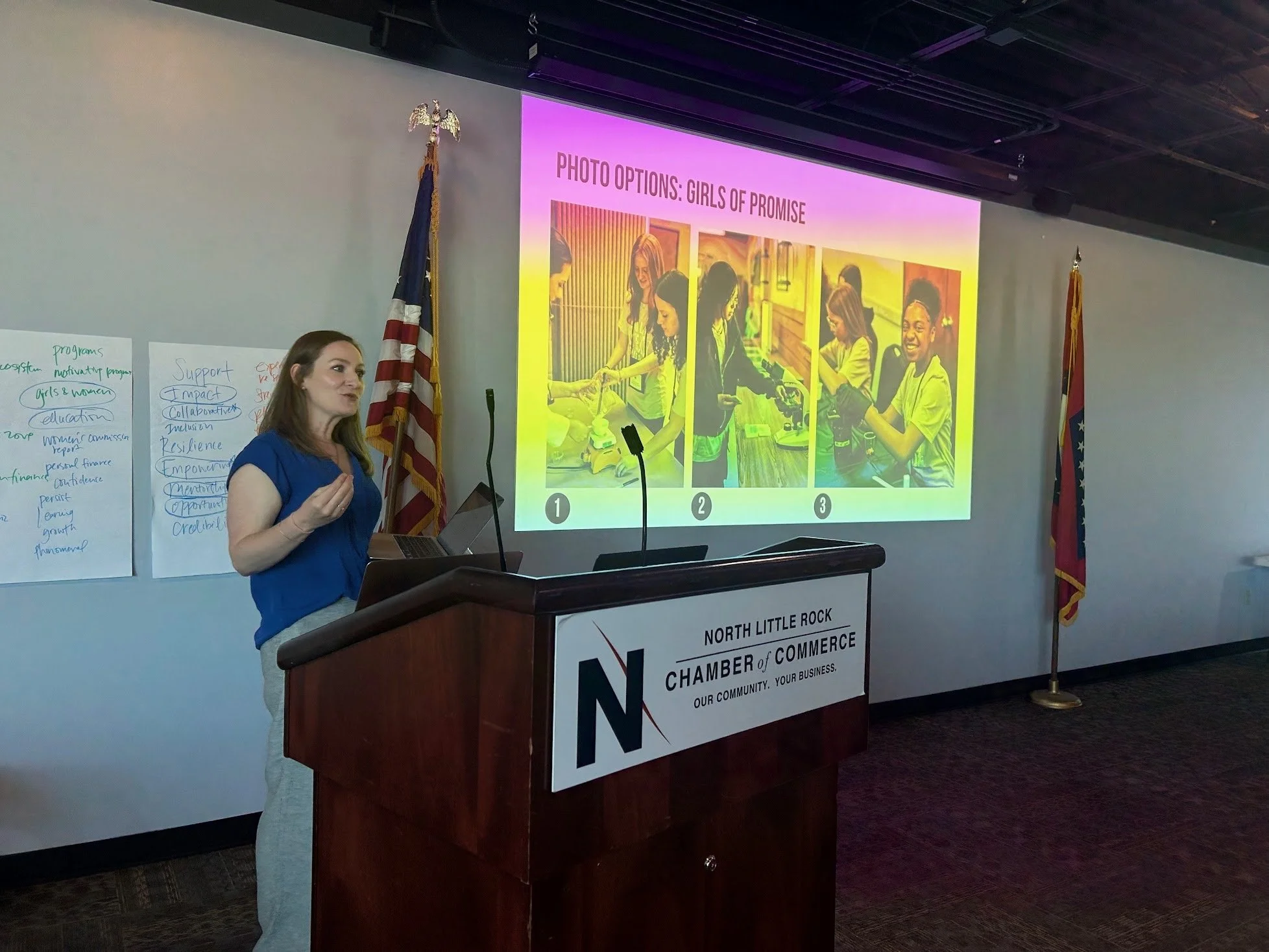 A woman giving a presentation at a wooden podium with a sign for North Little Rock Chamber of Commerce. She is facing a large screen showing a slide titled "Photo Options: Girls of Promise" with three images of girls working on projects. The American flag is on her left, and the Arkansas state flag is on her right.