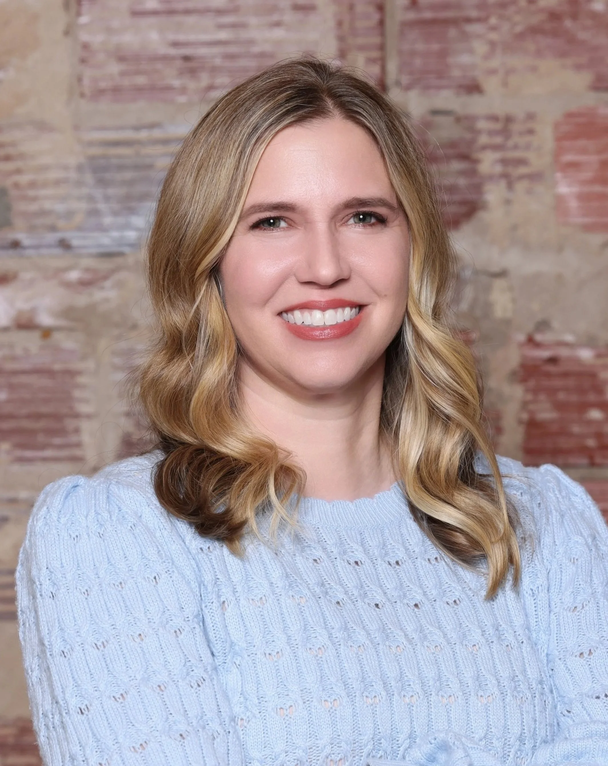 Portrait of a smiling woman with shoulder-length blonde hair, wearing a light blue knit sweater, standing in front of a brick wall.