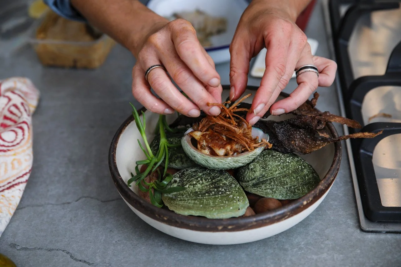 Person garnishing a bowl of oysters with shredded meat or seafood, surrounded by green succulents and other shellfish, on a gray surface.