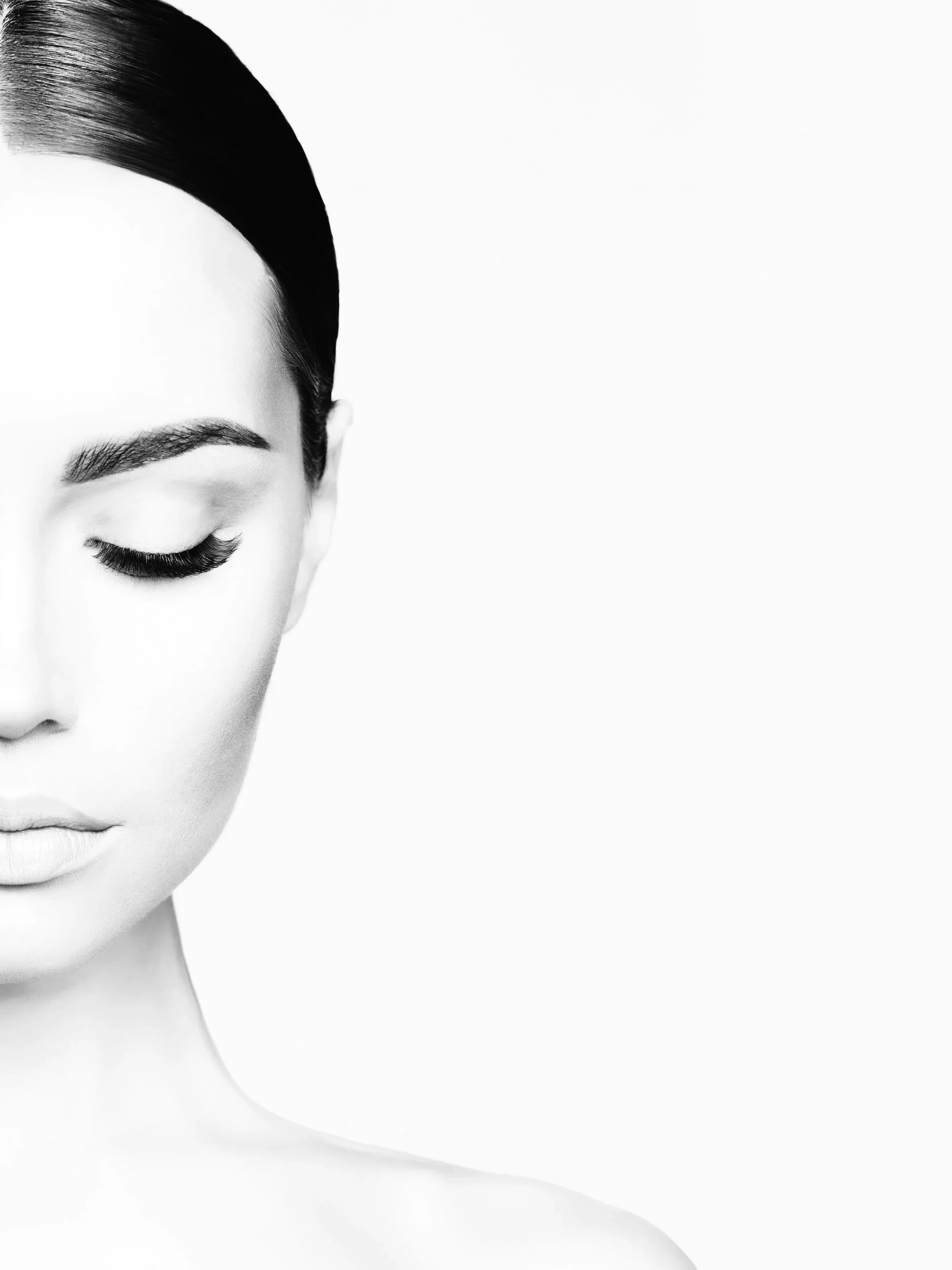 Black and white close-up of a woman's face with eyes closed, showing smooth skin, dark eyebrows, and long eyelashes, against a plain white background.
