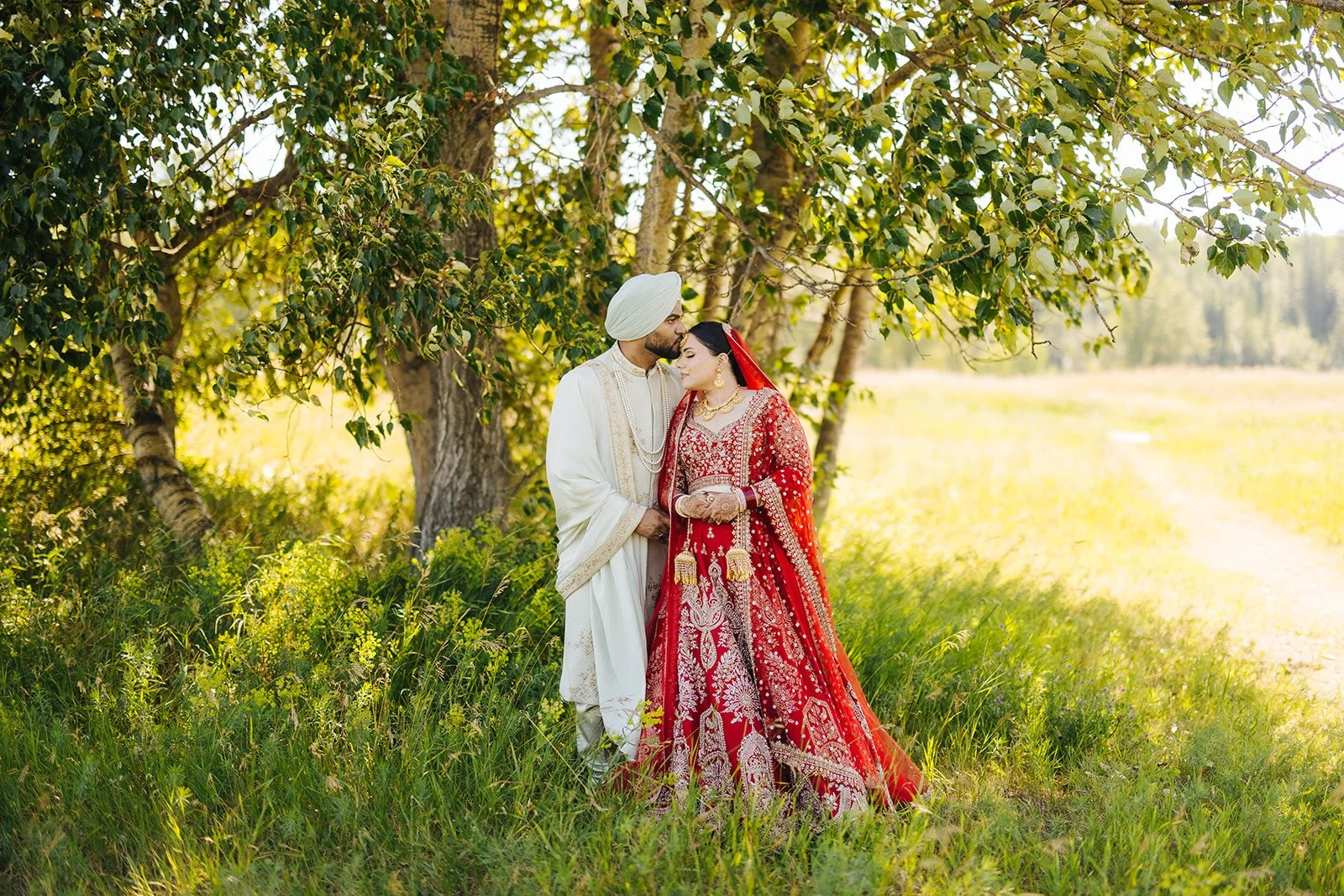 A couple dressed in traditional Indian wedding attire sharing a kiss in a sunlit forest.