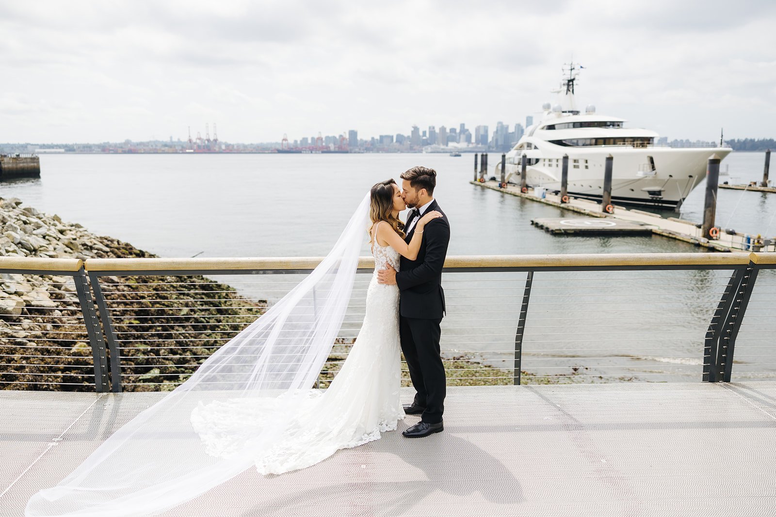 A bride and groom embracing and kissing on a waterfront deck with a yacht and city skyline in the background.