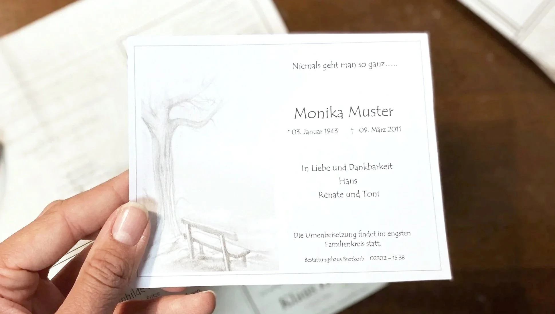 Eine Hand hält eine Beileidskarte mit einem Baum auf der linken Seite. Die Karte enthält den Namen Monika Muster, Geburtsdatum und Sterbedatum. Es ist ein Kondolenztext mit Danksagung