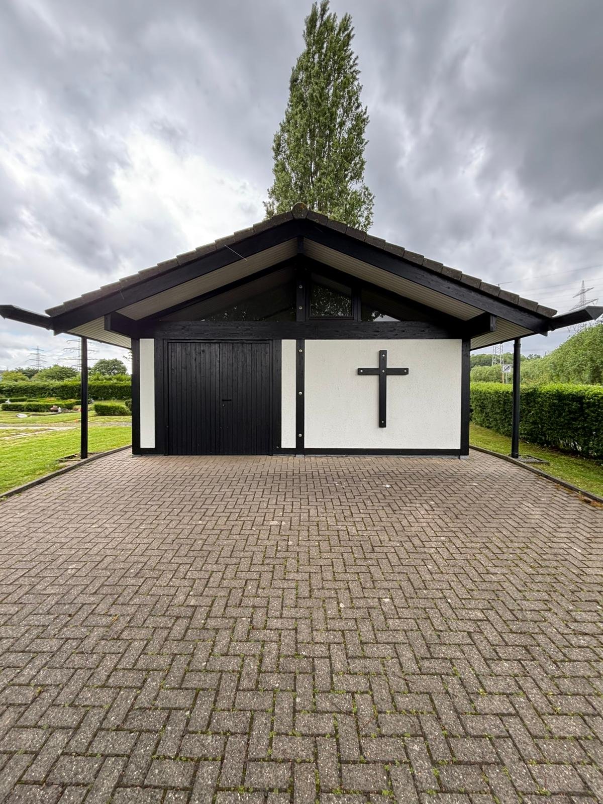 Ein kleines christliches Gotteshaus mit schwarzem Dach und weißen Wänden, auf einer gepflasterten Fläche, mit einem Holzkreuz an der Wand auf der rechten Seite, umgeben von Grünlandschaft und einem bewölkten Himmel.