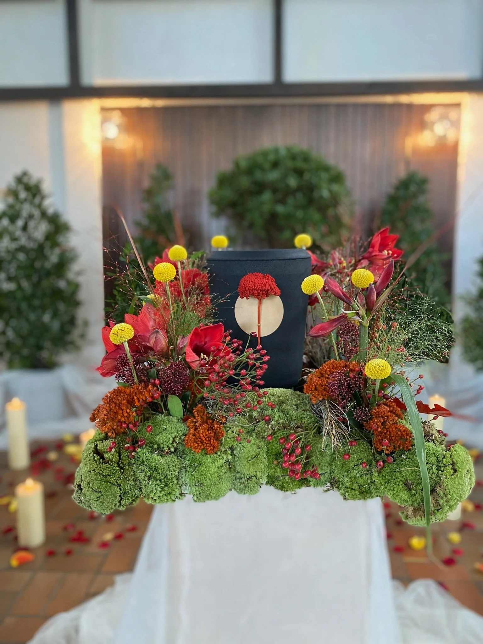 Kohleurne mit getrockneten roten Blumen in einem Blumenarrangement mit roten, gelben und orangefarbenen Blumen und Moos, auf einem weißen Tuch mit Kerzen im Hintergrund.