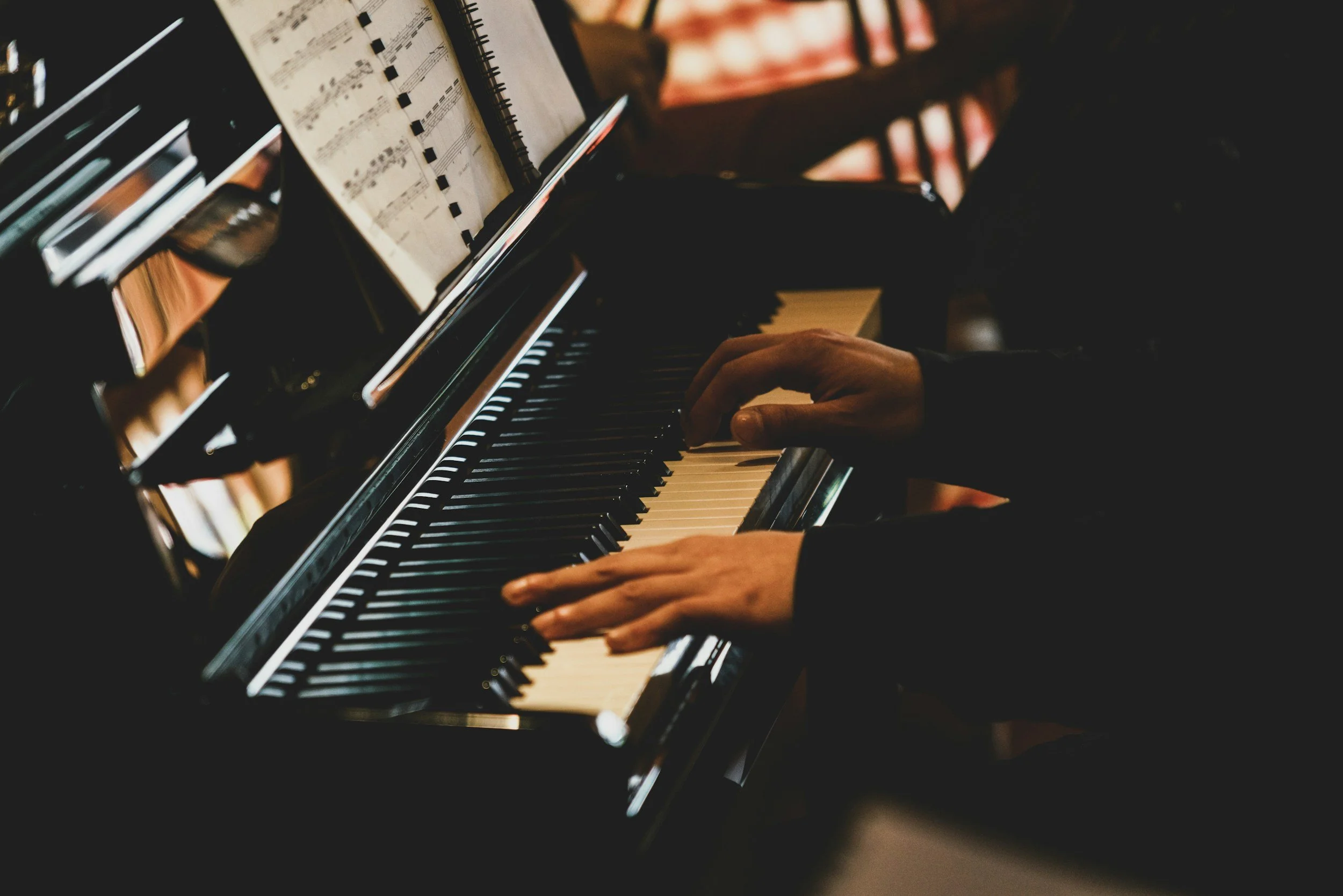 Ein Musiker spielt Klavier, wobei die Hände auf den Tasten liegen, und es liegen Notenblätter auf dem Notenständer. Das Bild ist düster beleuchtet.