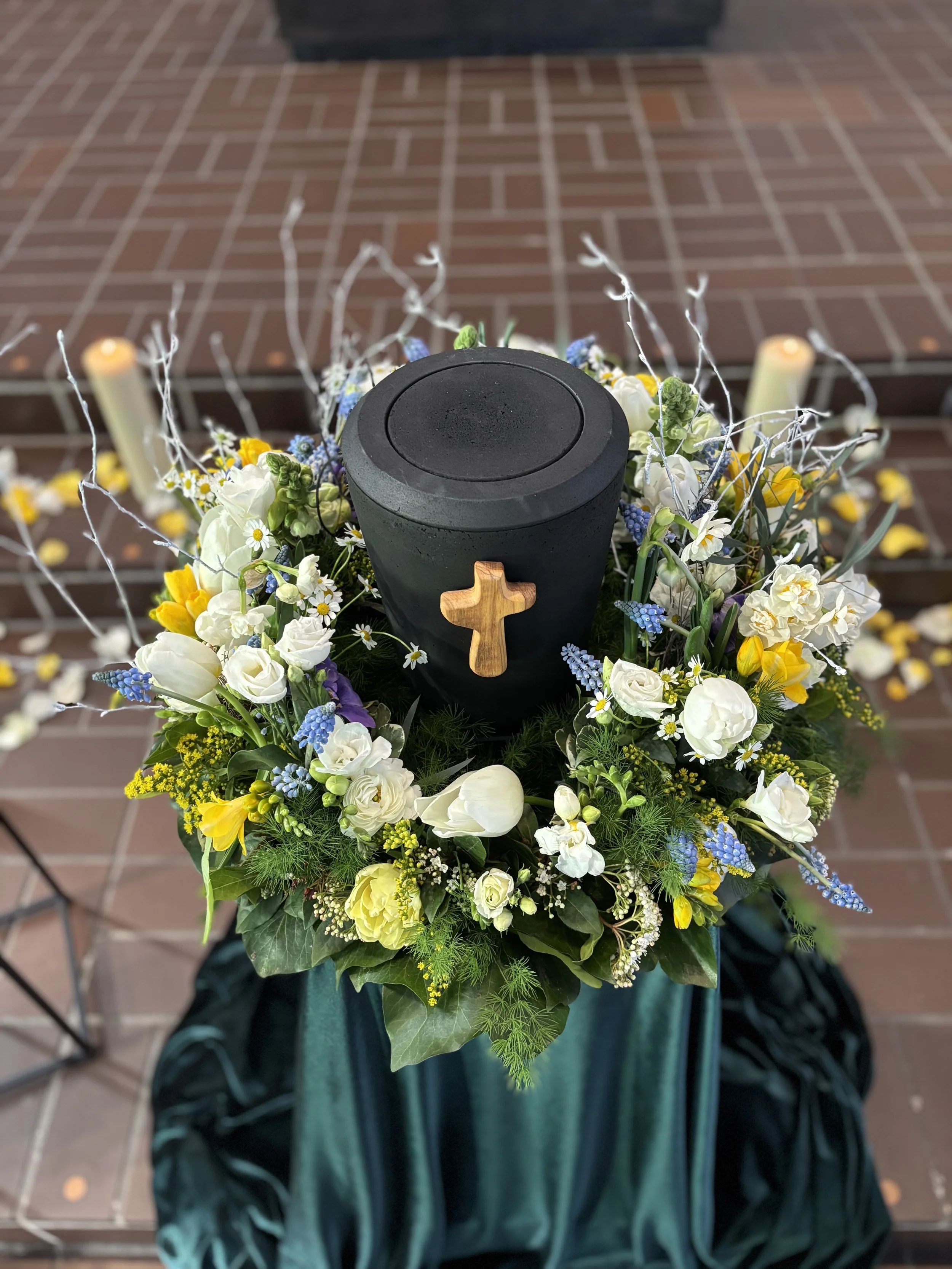 Kohleurne mit einem Kreuz aus Olivenholz in einem Blumenkranz mit weißen, gelben und blauen Blumen.