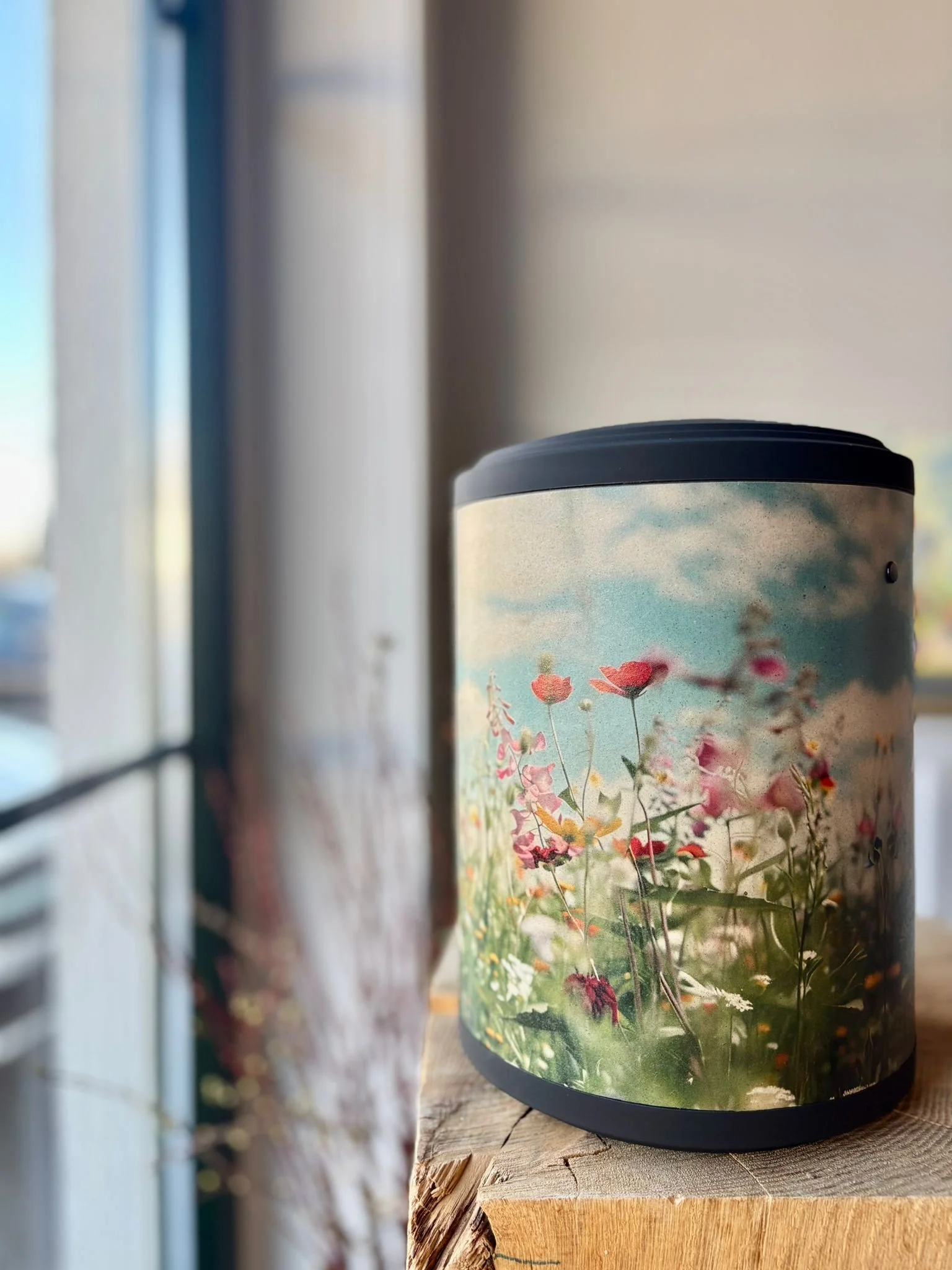 Urne aus Papier mit einer bunten Blumenwiese auf Holzblock neben Fenster