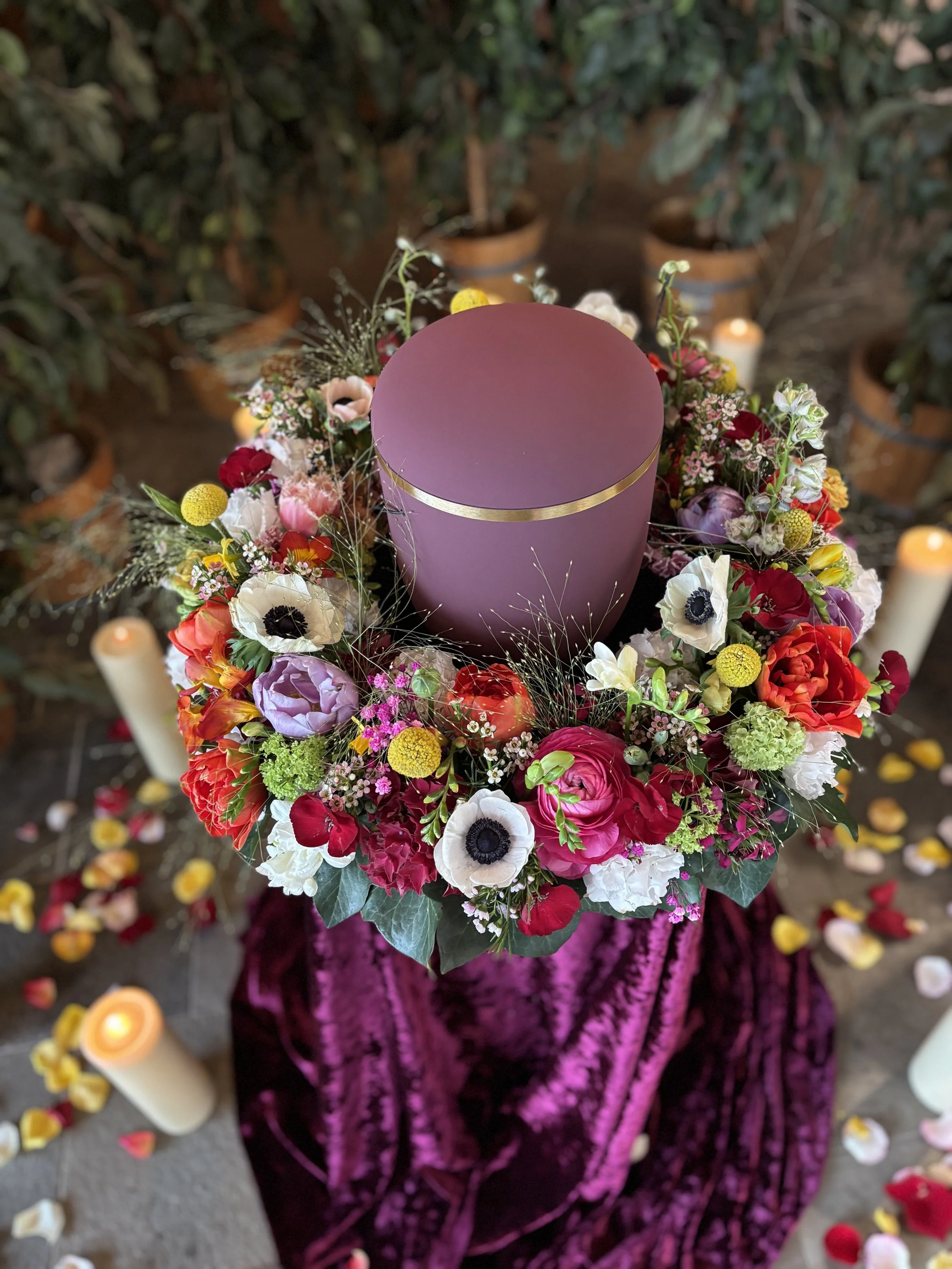 Runder Blumenkranz mit roten, weißen, lila und gelben Blumen, um einen pinkfarbenen Kerzenhalter mit Goldstreifen, auf einem violett-glänzenden Tuch, mit Kerzen und Blumenblättern auf dem Boden