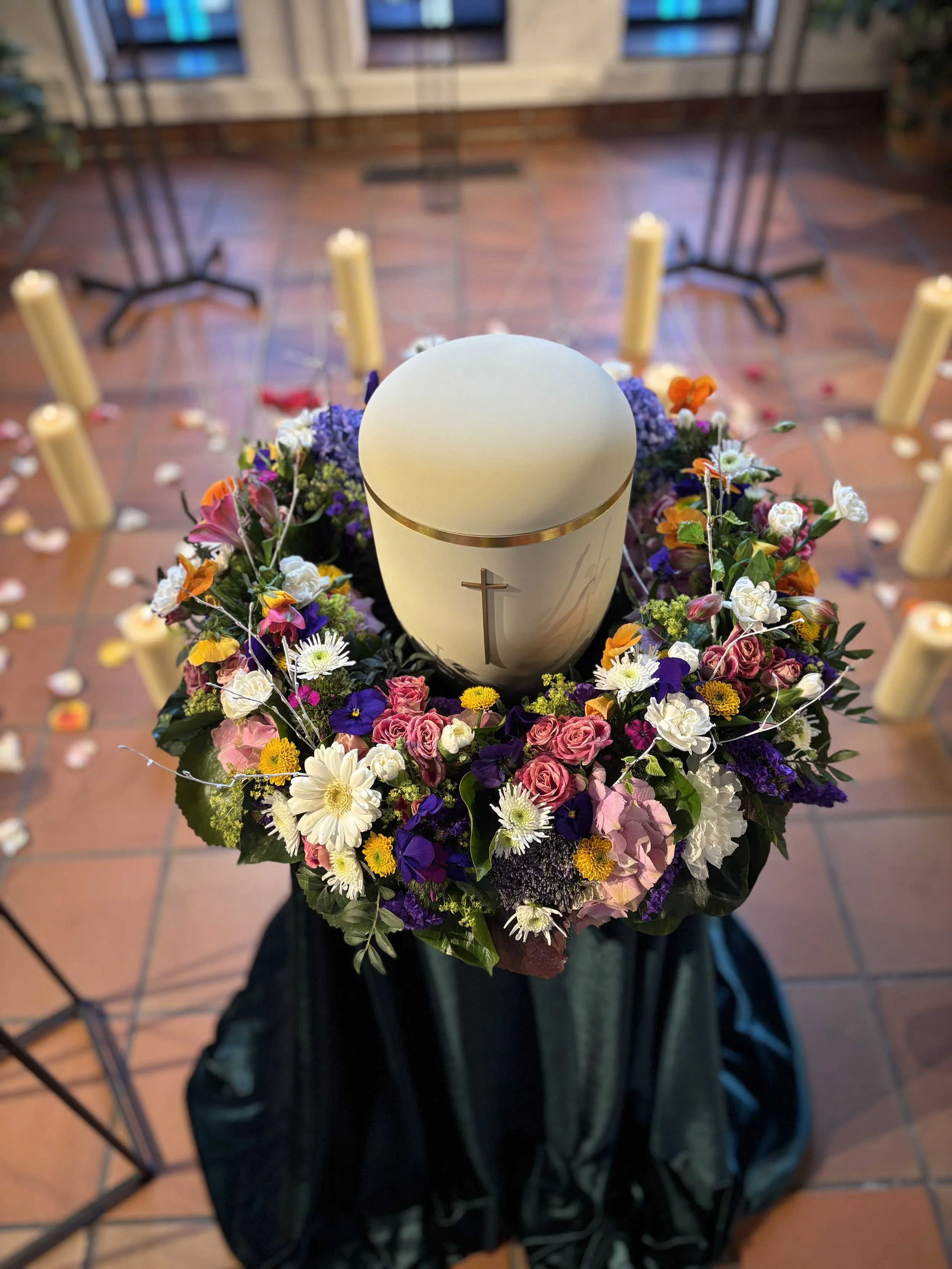 Weiße Urne mit goldener Bordüre inmitten eines bunten Blumenkranzes, umgeben von Kerzen in einer Kapelle.