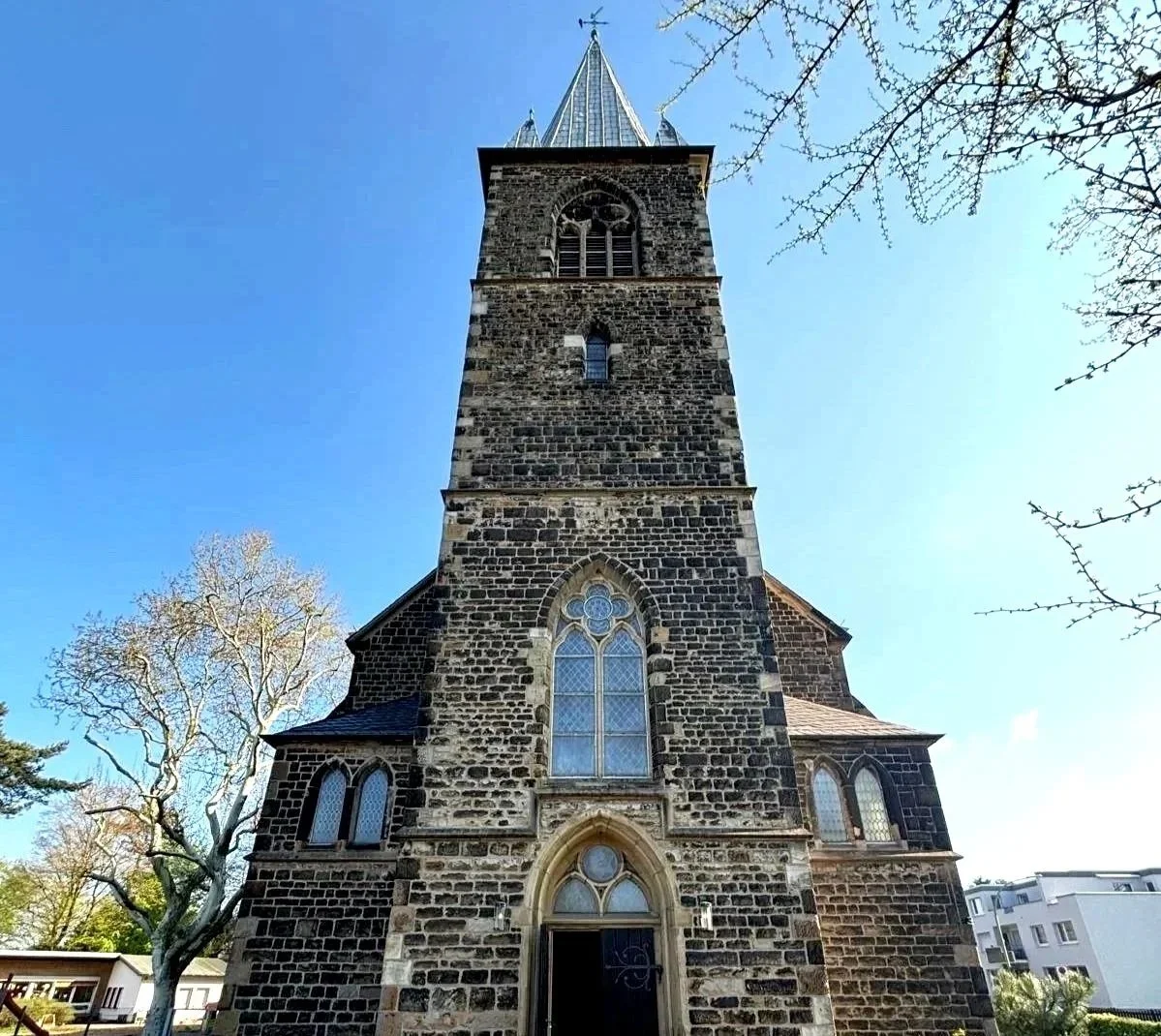 Ein gotischer Kirchturm aus dunklem Stein mit Fensteröffnungen, umgeben von Bäumen und modernen Gebäuden im Hintergrund der Trinitatis Gemeinde Witten Heven. Trauerhalle für den Friedhof Heven Steinhügel und Billerbeckstraße