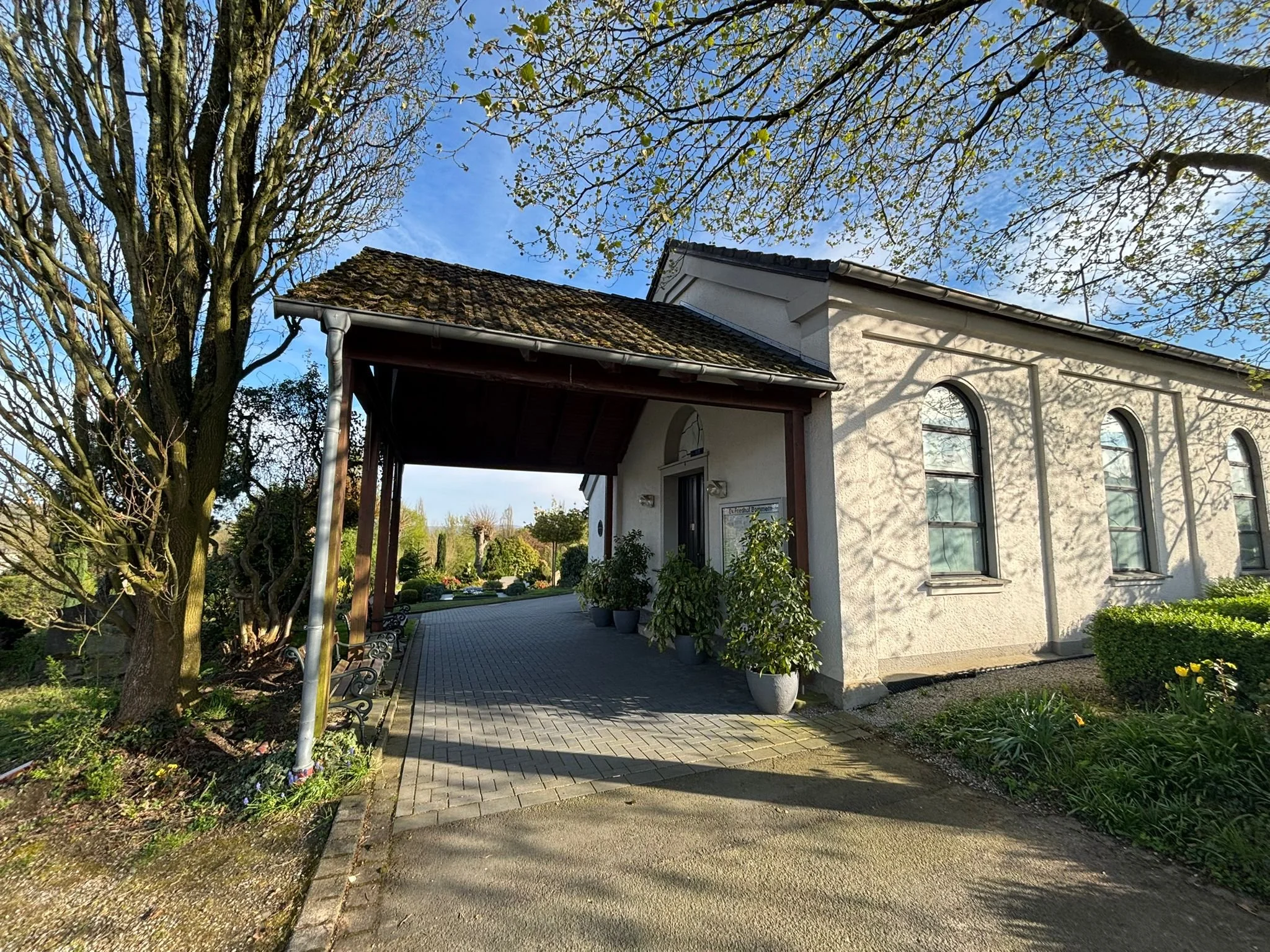 Trauerhalle des evangelischen Friedhof Witten-Bommern . Ein weißes Haus mit einem schrägen Dach und großen Fenstern, umgeben von einem Garten mit Bäumen, Sträuchern und einer gepflasterten Einfahrt, bei schönem Wetter an einem klaren Tag.