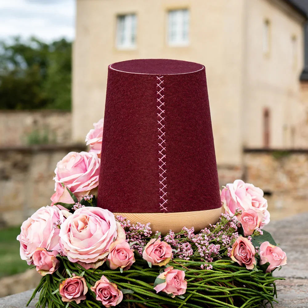 Eine Urne aus dunkelrotem Filz mit Holzsockel in der Mitte auf einem Blumenkranz aus rosa Rosen und kleinen lilafarbenen Blumen, im Hintergrund ein Haus, das im Freien steht.