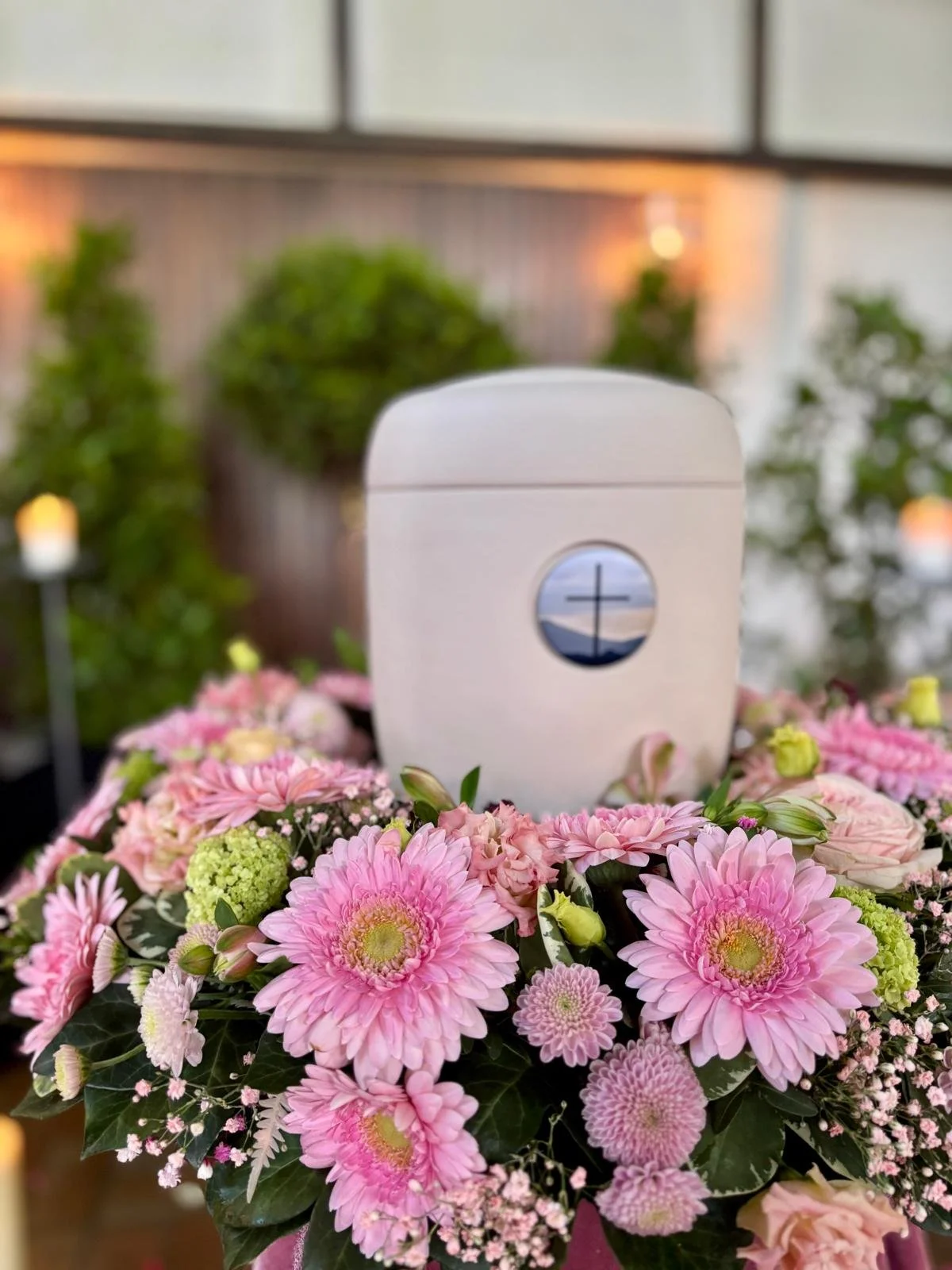 Kreuz auf einer weißen Urne auf einem Blumenkranz aus pinken, weißen und grünen Blumen, im Hintergrund Pflanzen und warme Beleuchtung.
