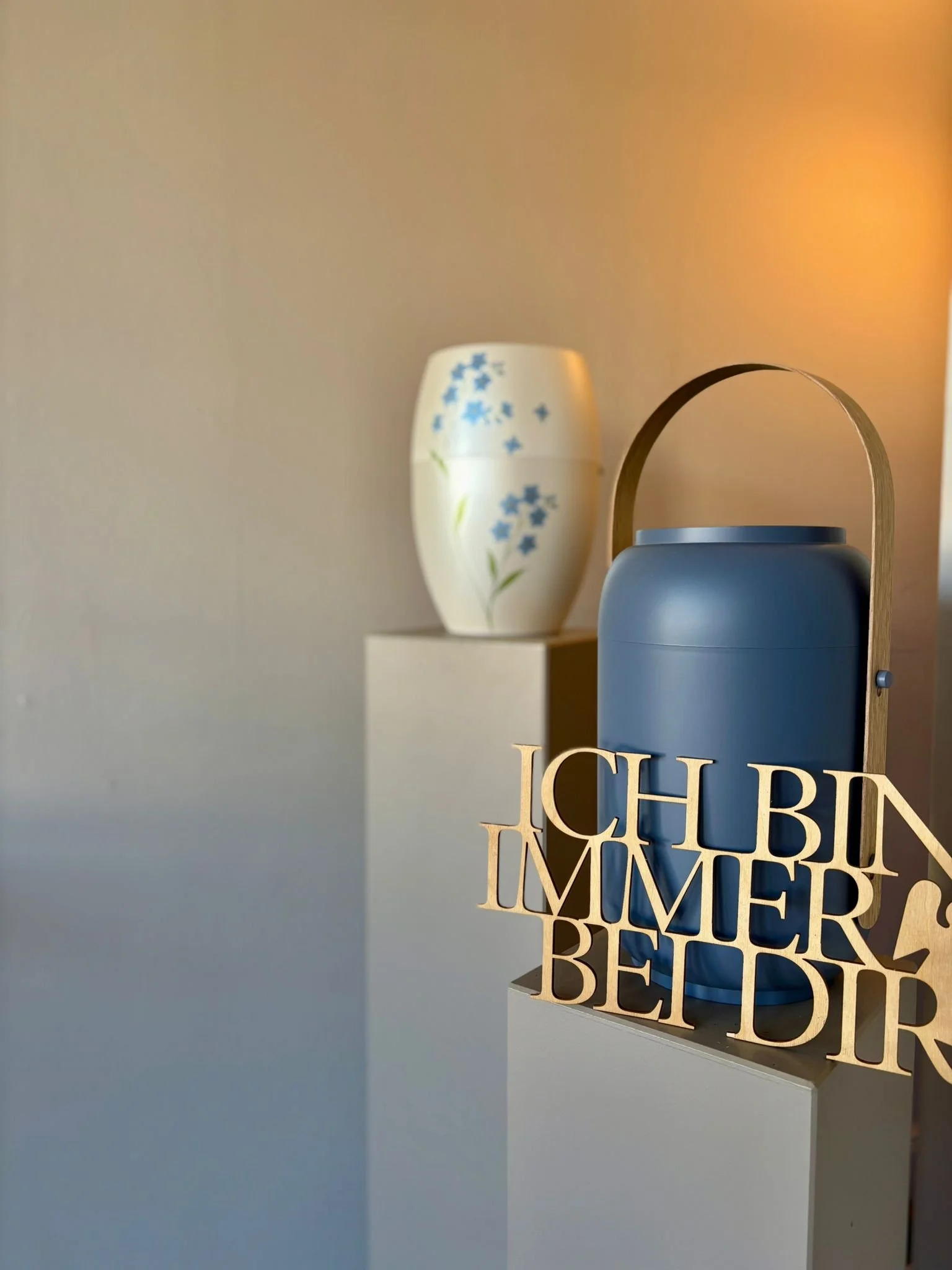 Eine Urne mit Vergissmeinnicht Motiv und eine moderne Urne in taubenblau mit Holzelement und ein Holzschriftzug, der sagt: 'ich bin immer bei dir'.