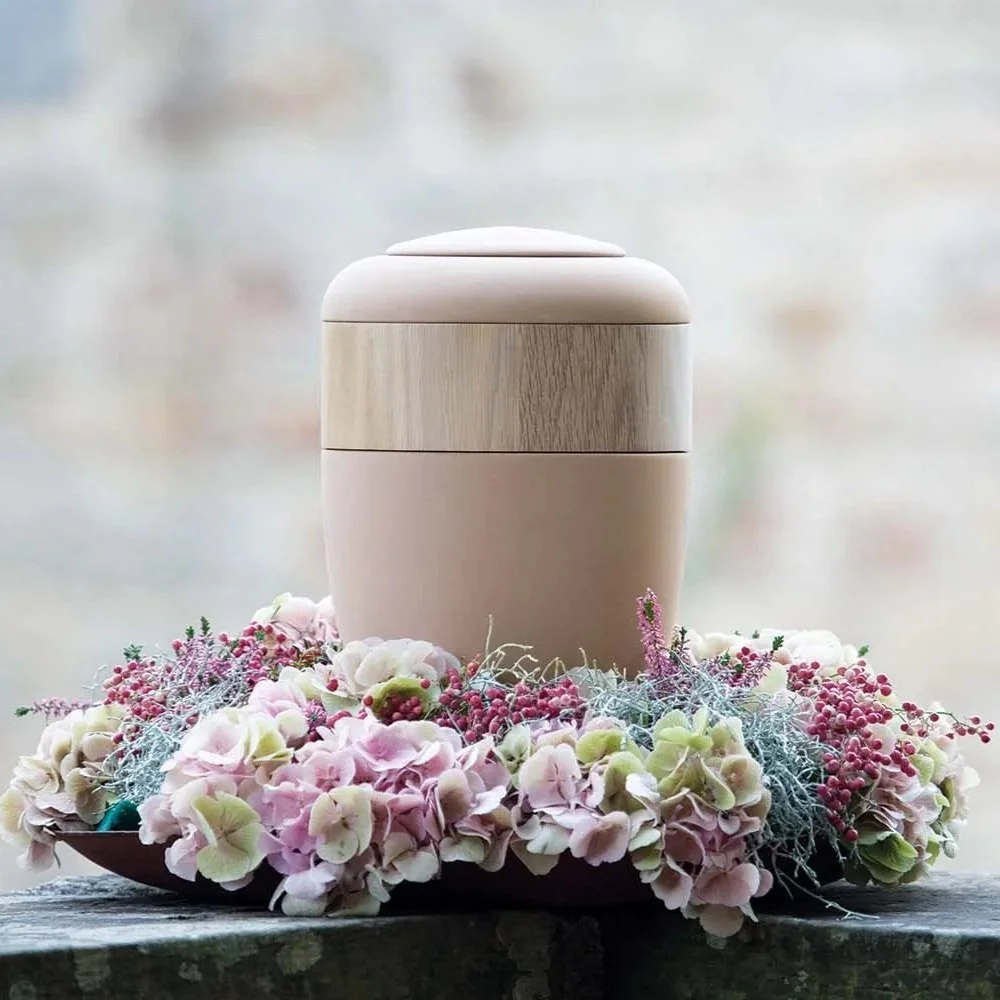 Ein runder, zartrosa Urne mit Ring aus hellem Holz auf einem Teller, umgeben von Blumen, vor einem verschwommenen hellen Hintergrund.