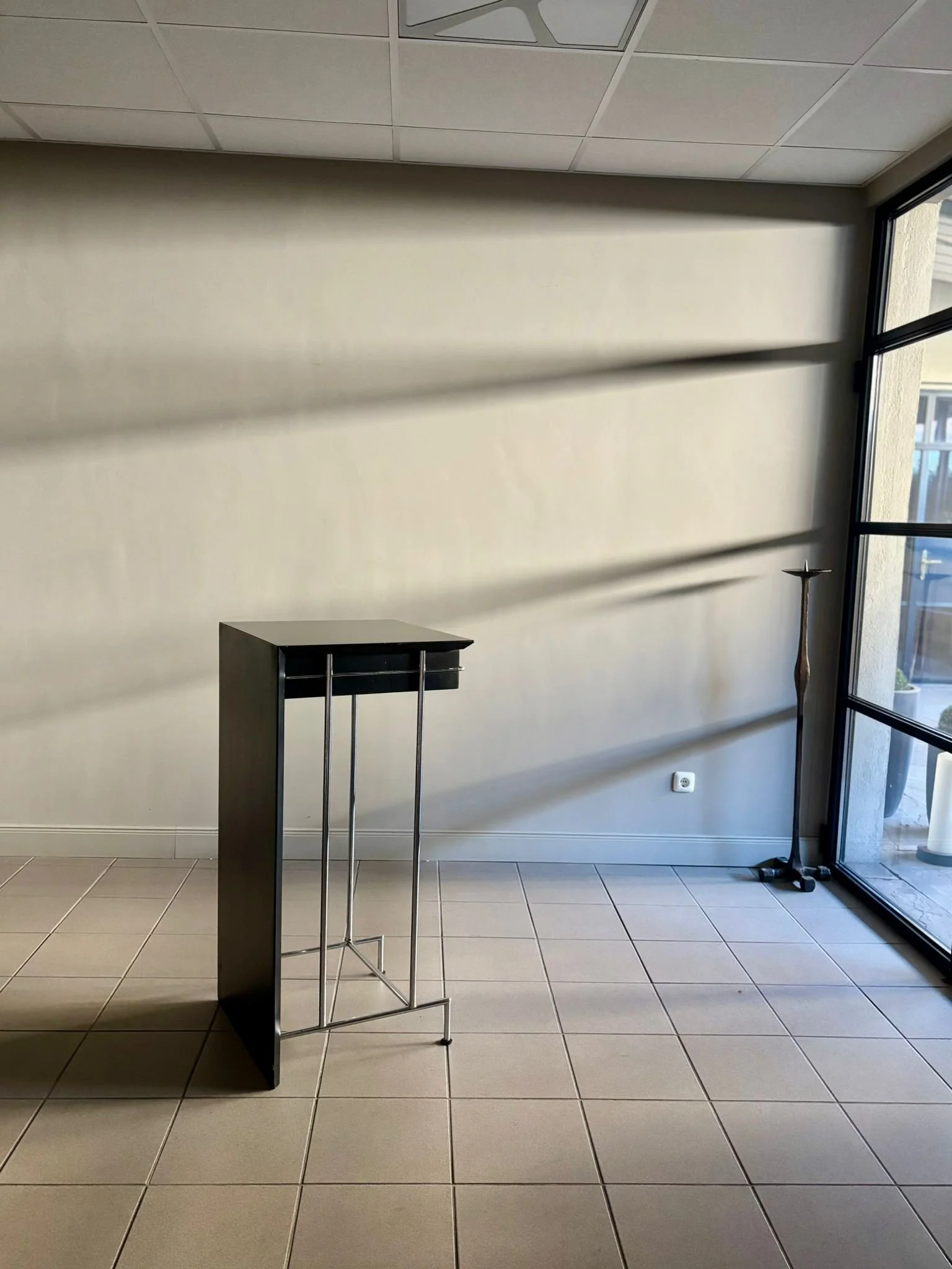 Ein leeres Zimmer mit Fliesenboden, einer großen Fensterfront auf der rechten Seite, einer schwarzen Ablage mit Metallstangen in der Mitte, einer dekorativen Stehleuchte in der Ecke rechts, und einer weißen Wand im Hintergrund.