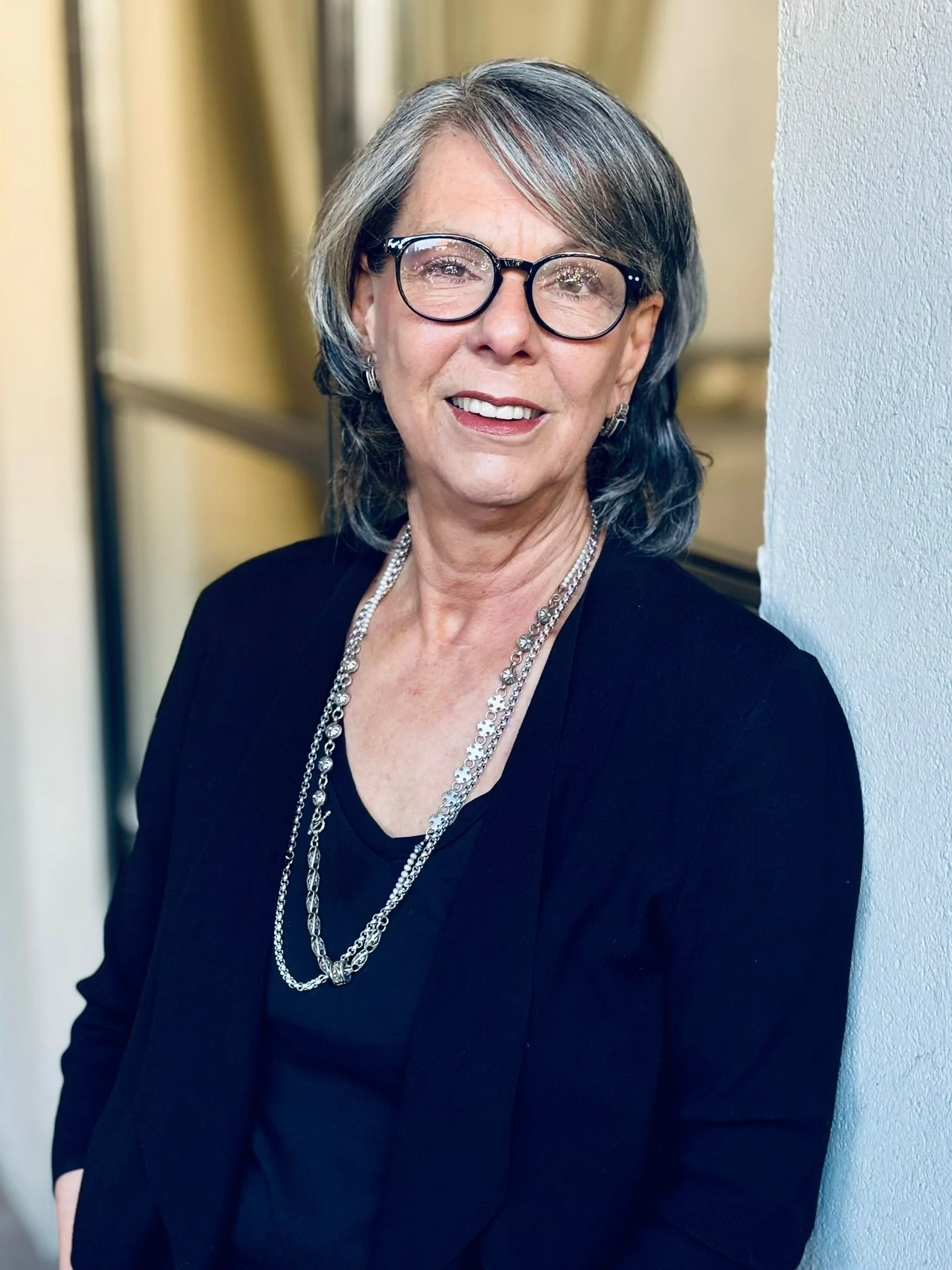 Eine Frau mit Brille, grauen Haaren und schwarzer Kleidung steht neben einer Wand, lächelt und trägt mehrere silberne Halsketten.