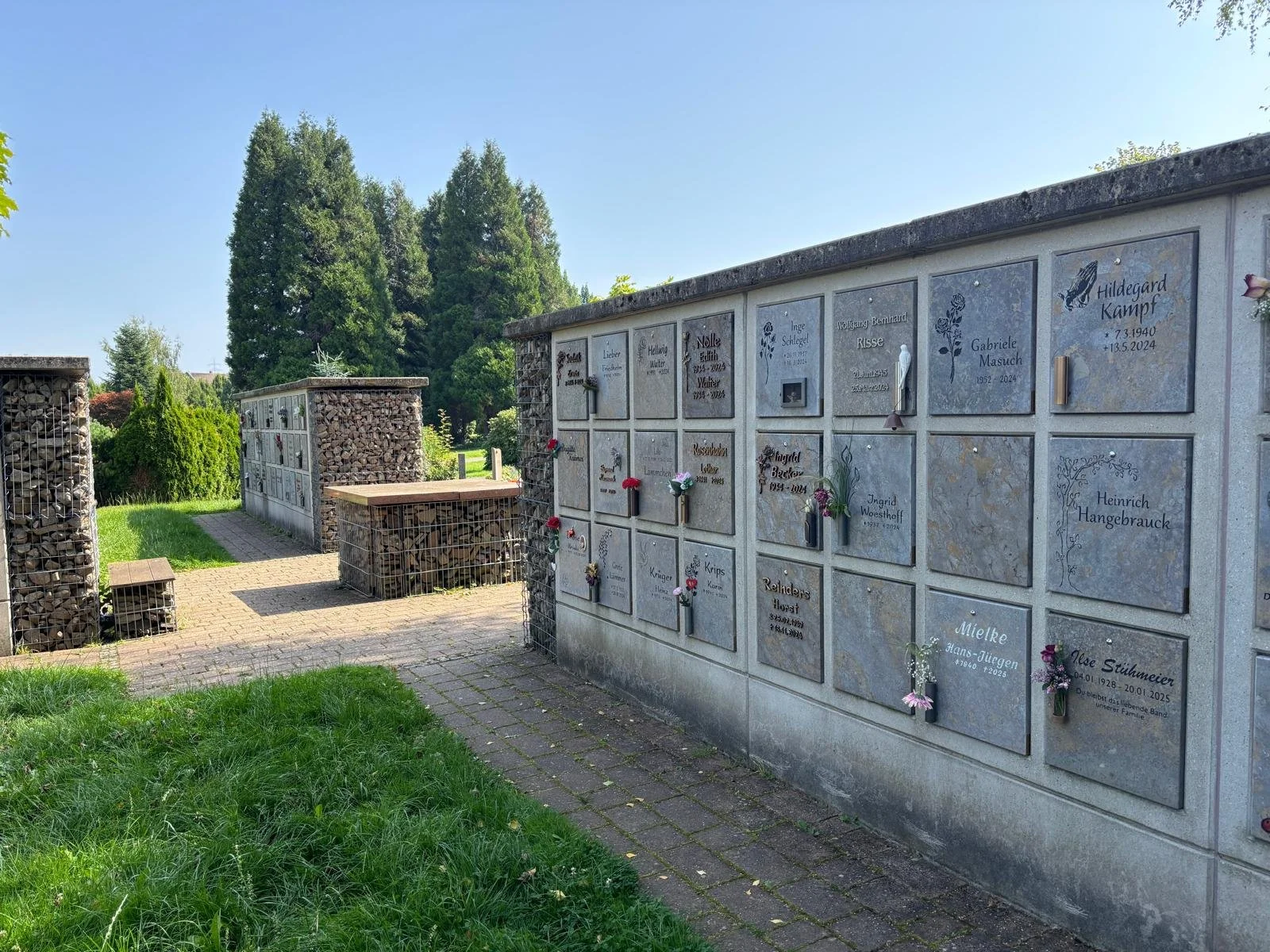 Kolumbarium auf dem Ev. Friedhof Pferdebachstraße in Witten. Dekorierte Gedenktafeln auf einem Friedhof mit Blumen, im Hintergrund Bäume und blauer Himmel.
