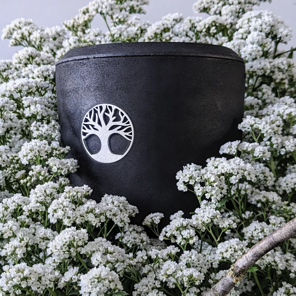 Schwarze Urne aus gepresster Kohle, umgeben von weißen Blumen, mit einem silbernen Baum-Logo.