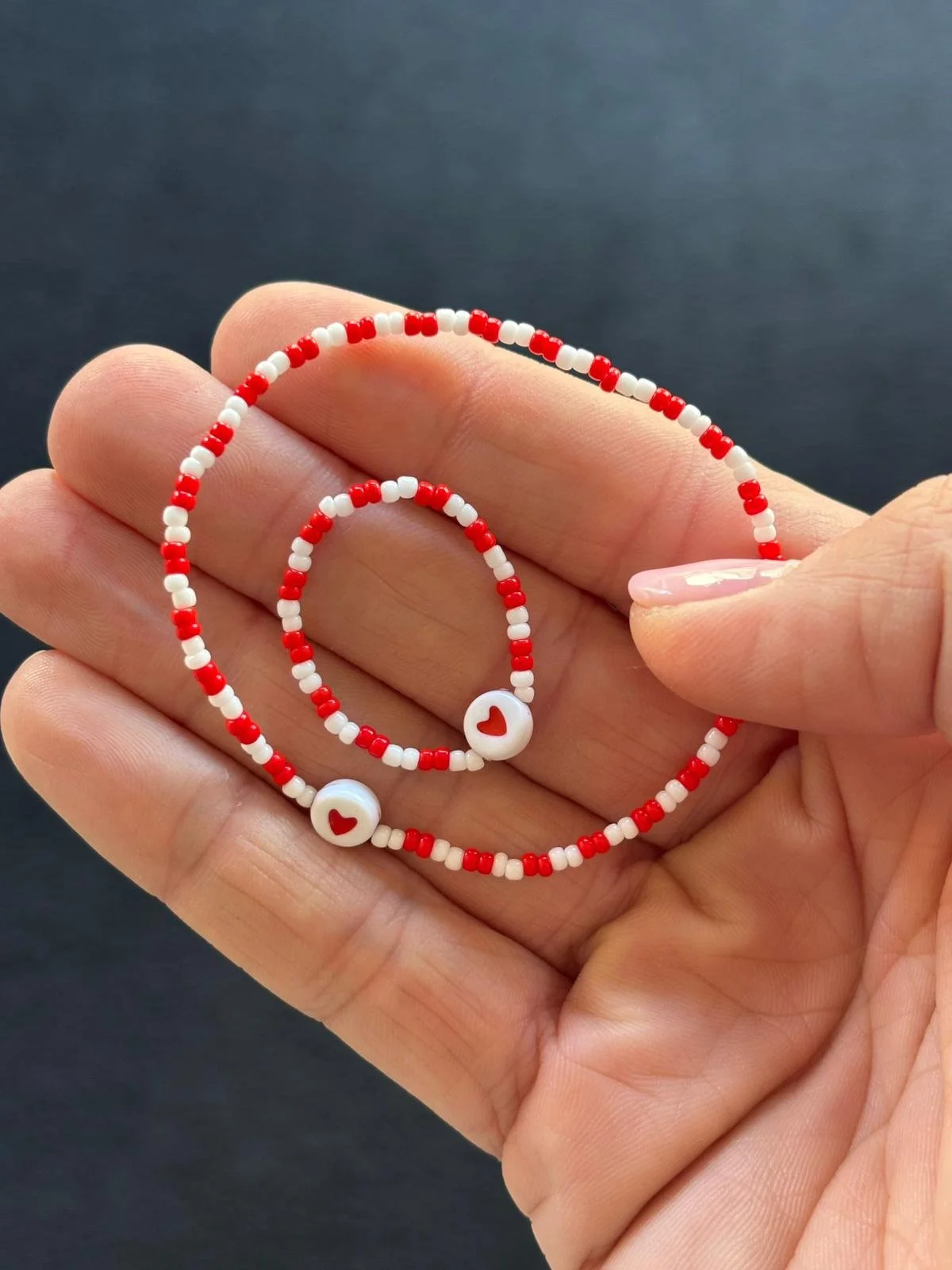 Hand hält zwei rot und weiß geknüpfte Armbänder mit Herzmotiven aus kleinen Perlen. Erinnerungsstücke für Sternenkinder und Frühchen von Herzenssache E.V.