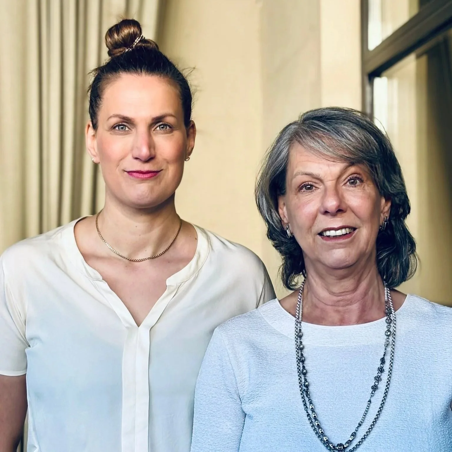 Zwei Frauen stehen nebeneinander vor einem hellen Hintergrund. Die junge Frau auf der linken trägt ein weißes Hemd und eine goldene Halskette, die ältere Frau auf der rechten trägt ein weißes T-Shirt mit mehreren silbernen Halsketten.