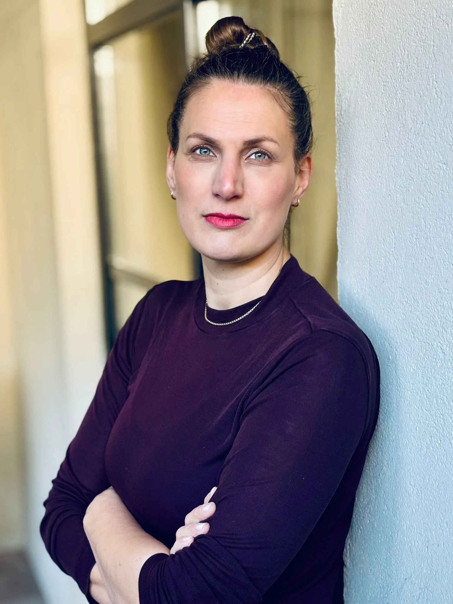 Porträt einer Frau mit blauen Augen, braunem Haar in einem hohen Dutt, dunkler Bluse, vor weißer Wand, Arme verschränkt.