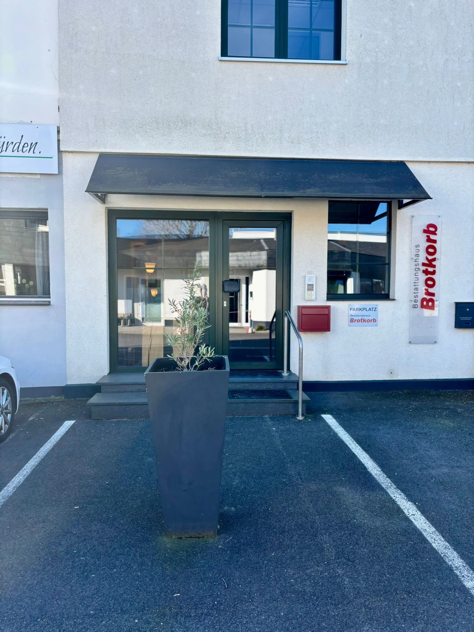 Eingang eines Gebäudes, Pflanzen im Vordergrund, Parkplätze vor dem Gebäude, Beschriftung 'Bestattungshaus Brotkorb' an der Wand.