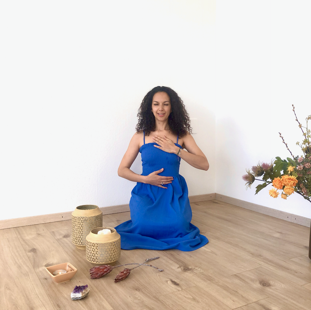 Person meditating in a blue dress with hand on chest and stomach, surrounded by meditation items like candles and flowers.