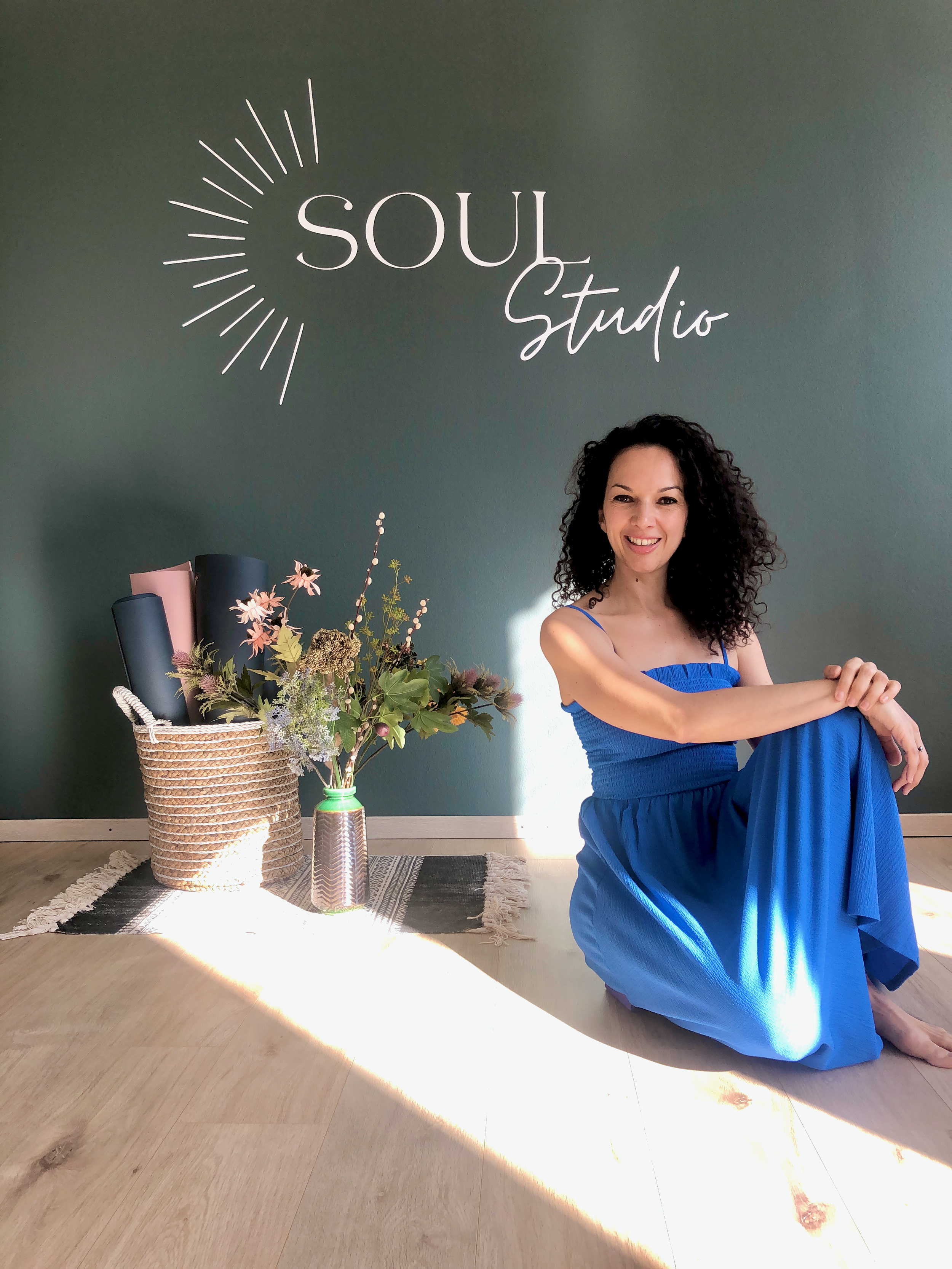 Woman in a blue dress sitting on the floor of a studio with a green wall and "Soul Studio" text, next to a basket containing yoga mats and flowers.