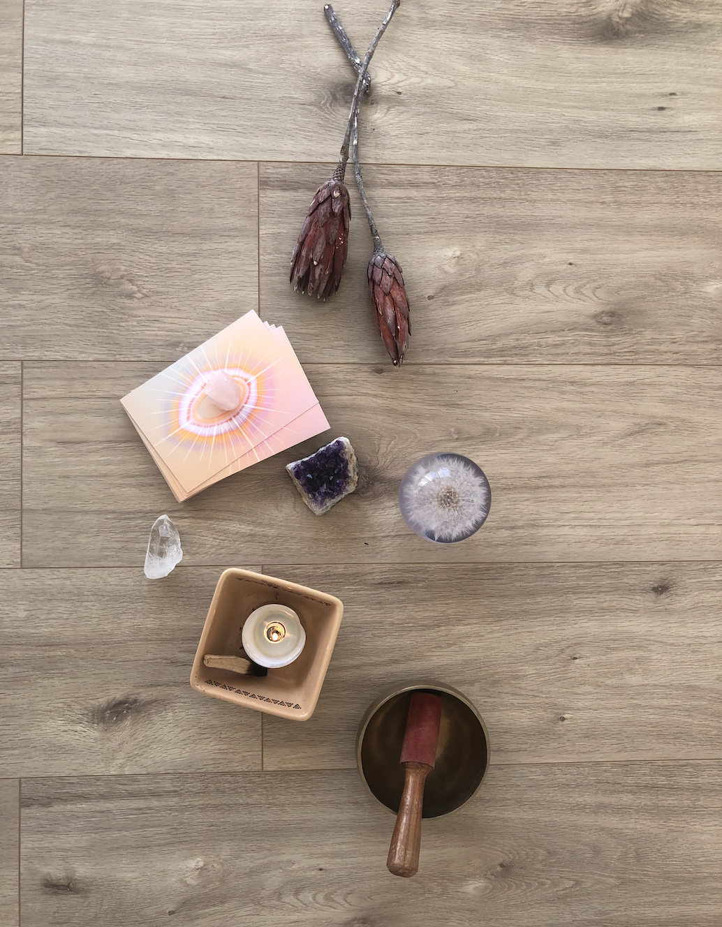 Flat lay of spiritual and meditation items on a wooden floor, including dried flowers, a set of cards with a planet design, amethyst and clear quartz crystals, a candle in a clay holder, and a singing bowl with a mallet.