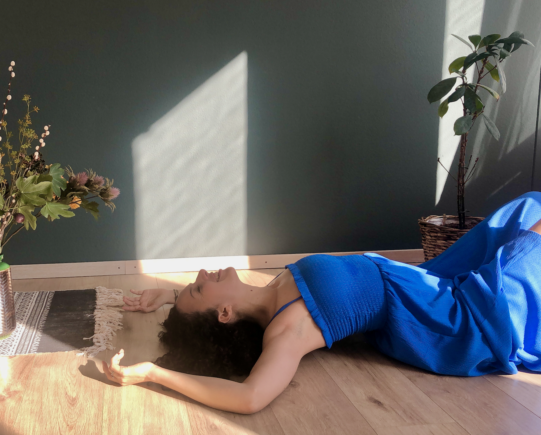 Woman in blue dress lying on floor near plants and woven mat, with sunlight on face, against a dark wall.