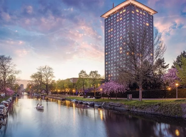 A cityscape at dusk with a river reflecting the sky and lights, a tall modern building with many windows, and trees with pink blossoms along the riverbank.