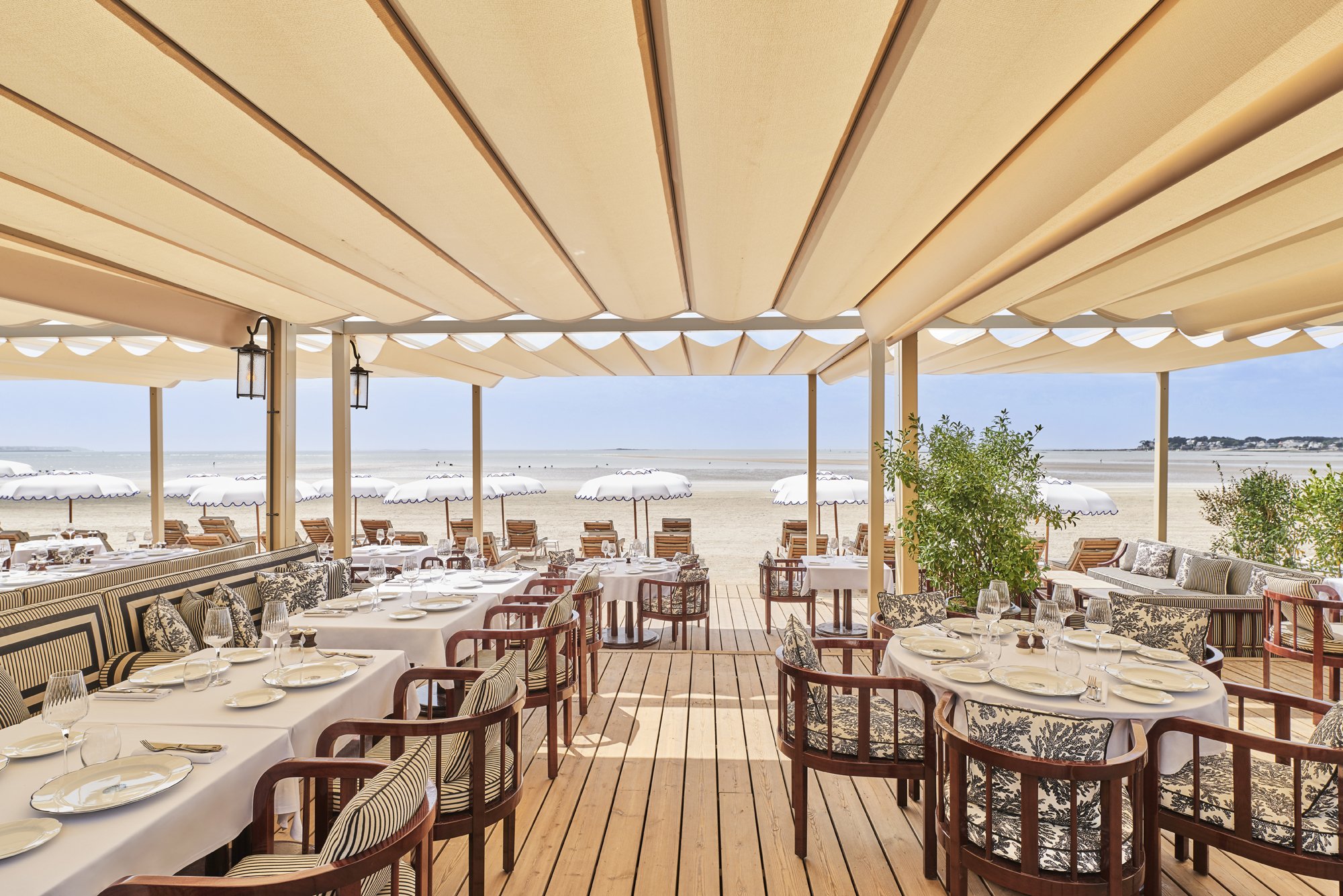 Beachside restaurant with neatly arranged tables, white tablecloths, and chairs, sheltered by beige awnings, overlooking the ocean with umbrellas and a sandy shore in the background.