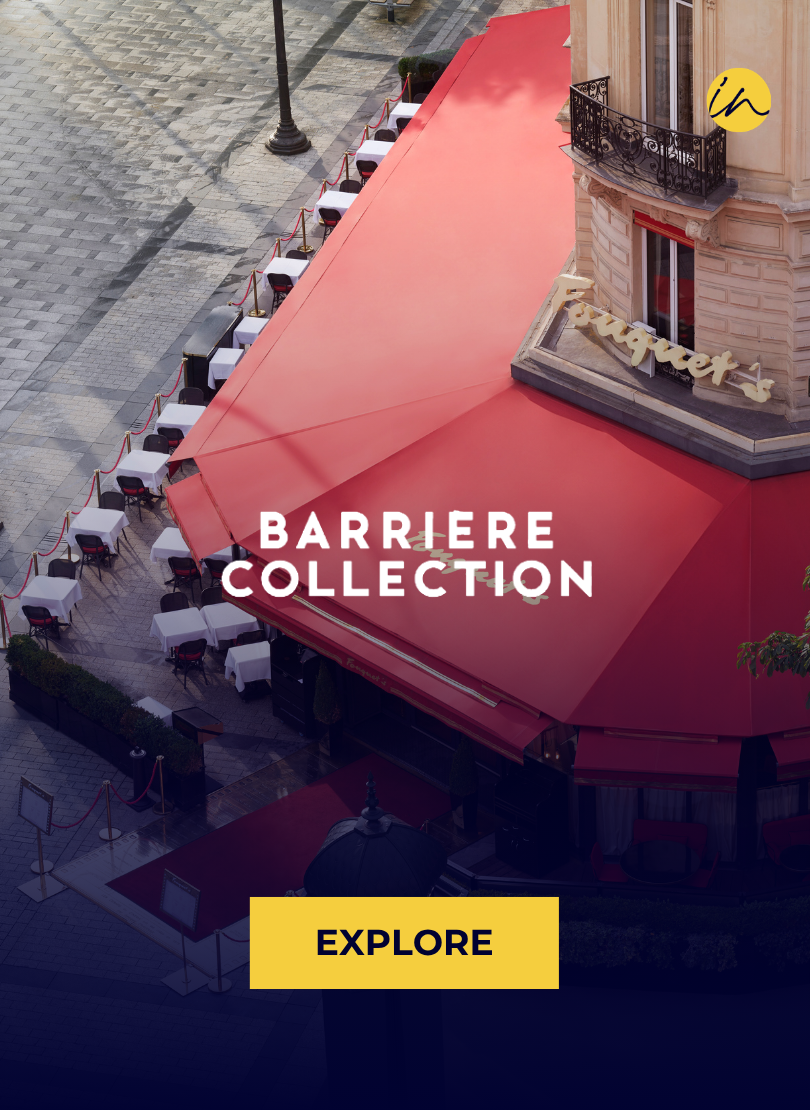 Empty outdoor restaurant patio with white tablecloth-covered tables, chairs, and red rope barriers, adjacent to a building with a pink awning, on a paved city street.