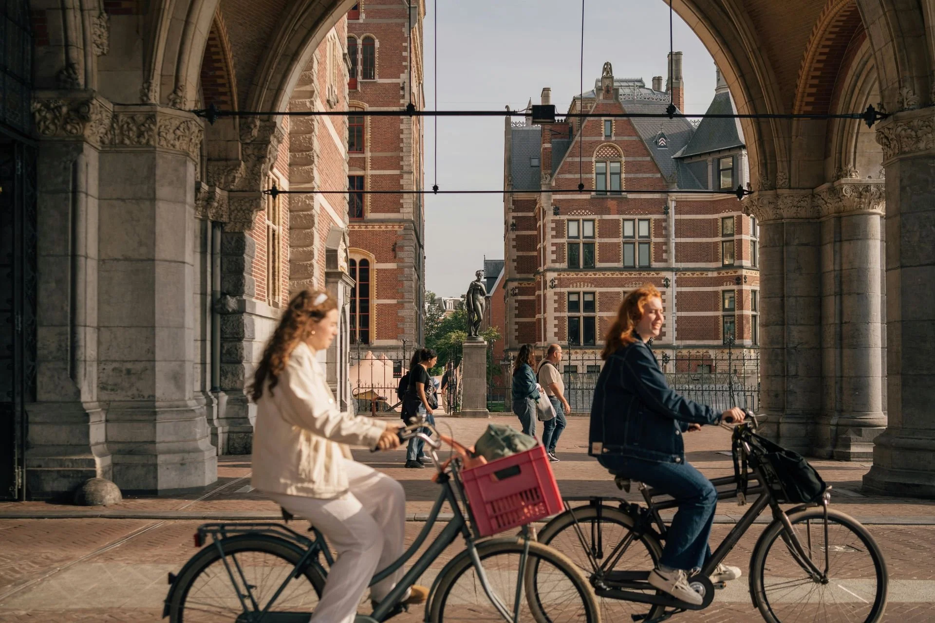 People riding bicycles beneath a stone archway in front of a historic brick building with ornate architectural details.