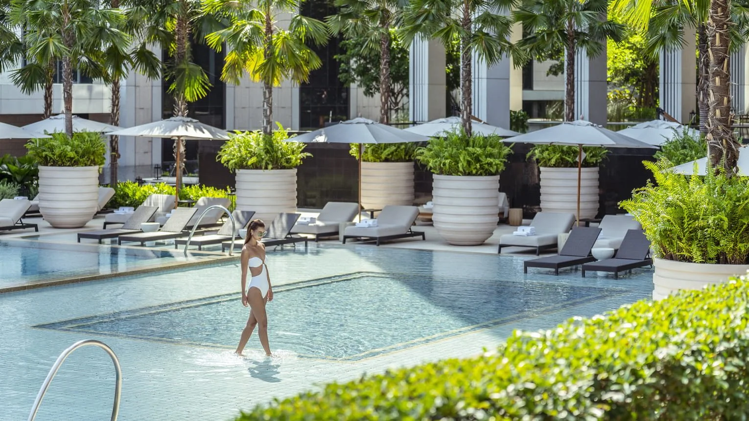 Woman wearing a white bikini walking in a swimming pool surrounded by pool chairs, umbrellas, and large potted plants.