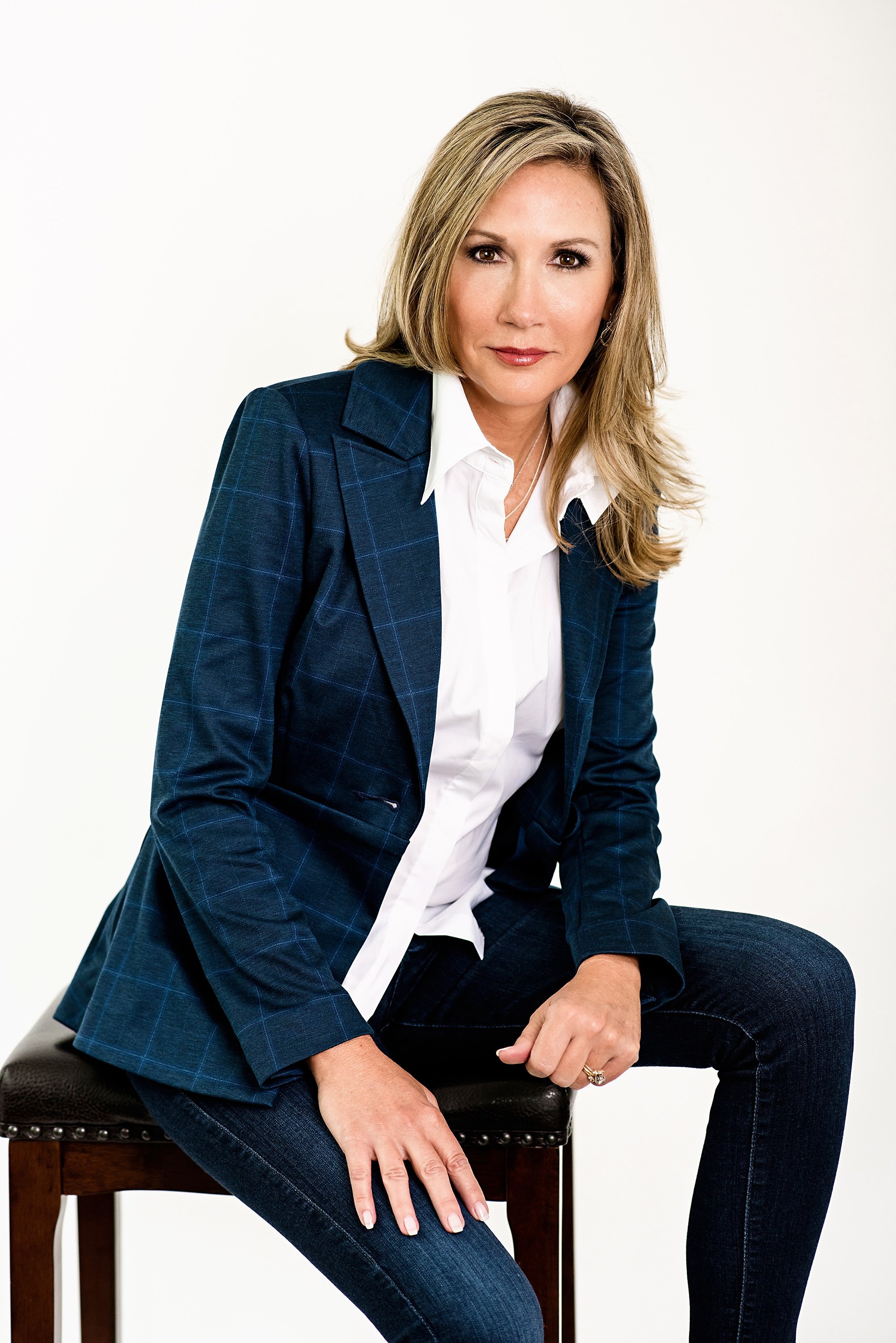 A woman with shoulder-length blonde hair, wearing a dark blue blazer with a subtle checkered pattern, white shirt, and dark jeans, sitting on a wooden stool against a plain white background.