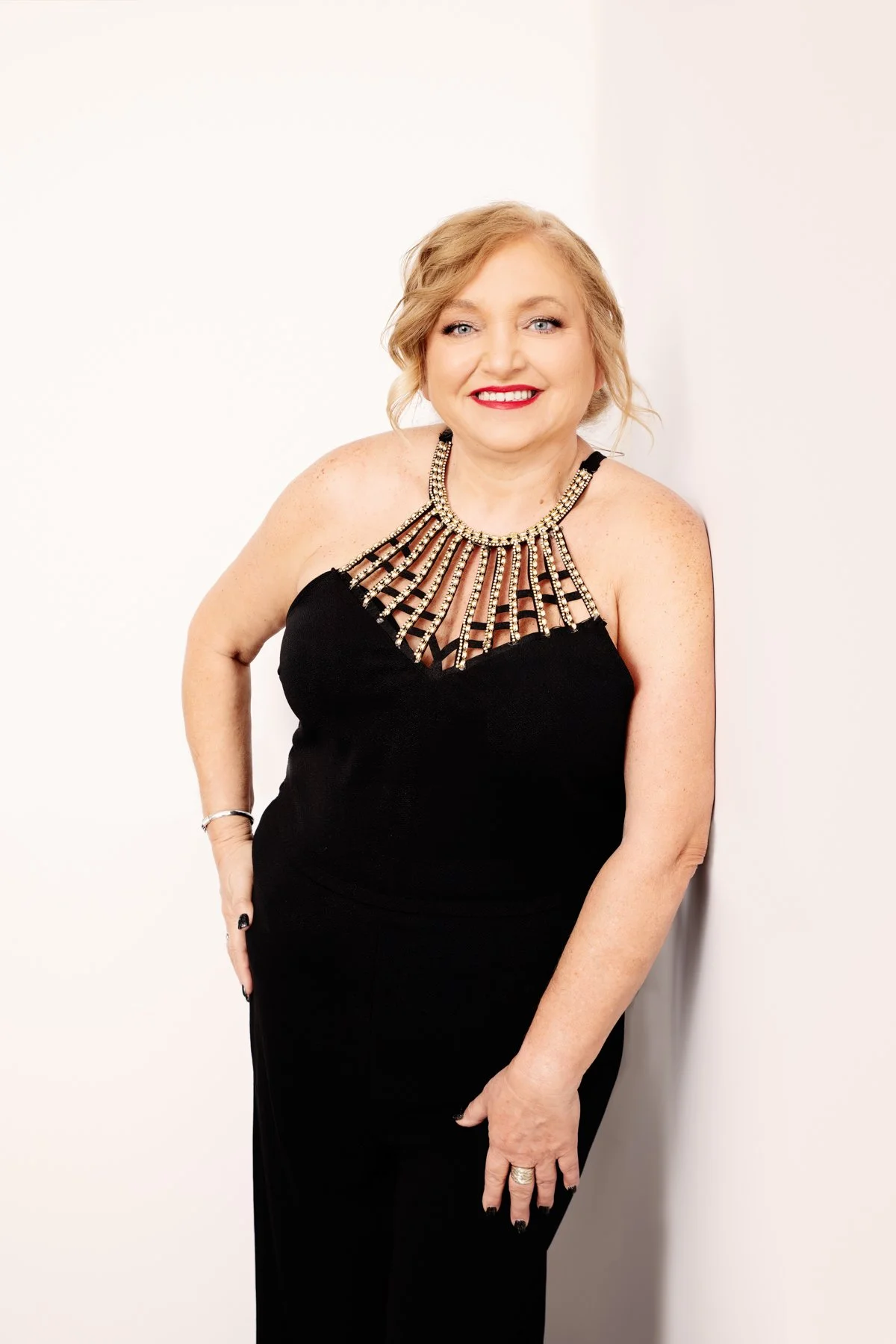 Woman with blonde hair in a black dress with an ornate, beaded neckline, smiling and standing against a light-colored background.