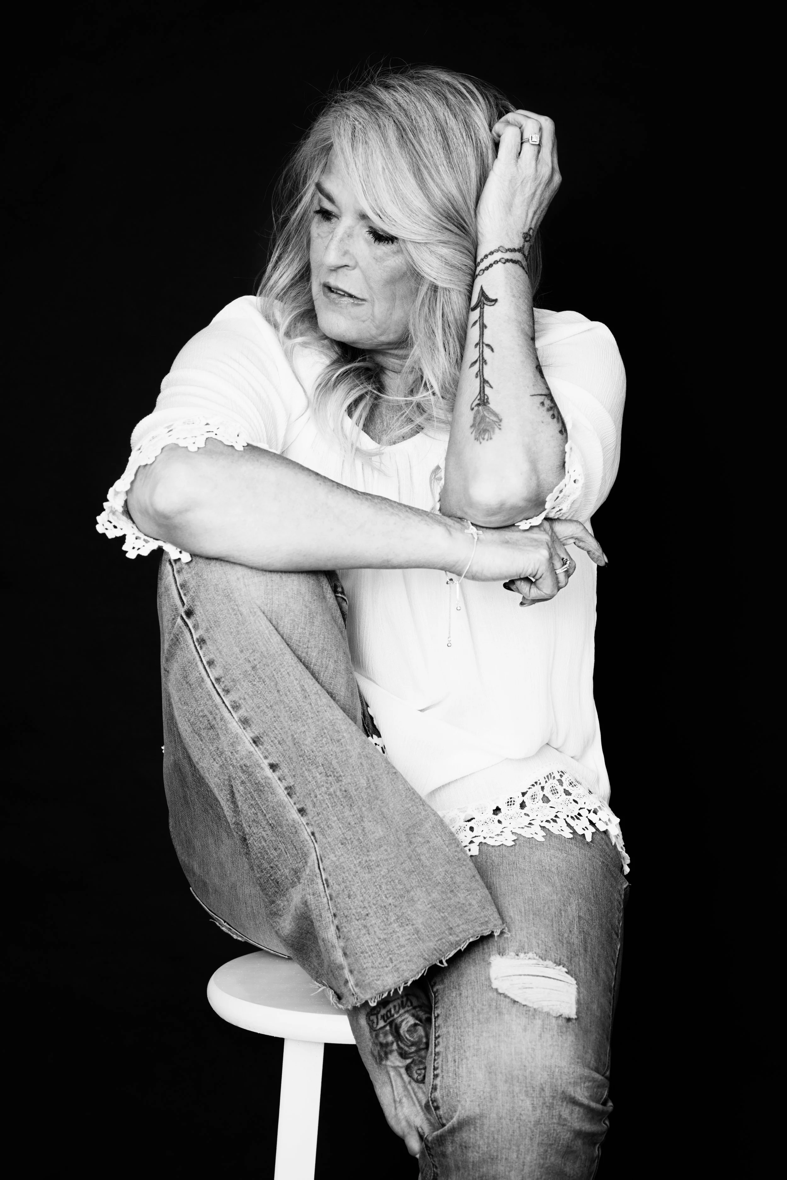 Black and white portrait of a woman with blonde hair, wearing a white top with lace details and ripped jeans, sitting on a stool against a dark background. She has tattoos on her arm and appears thoughtful, with her hand in her hair.