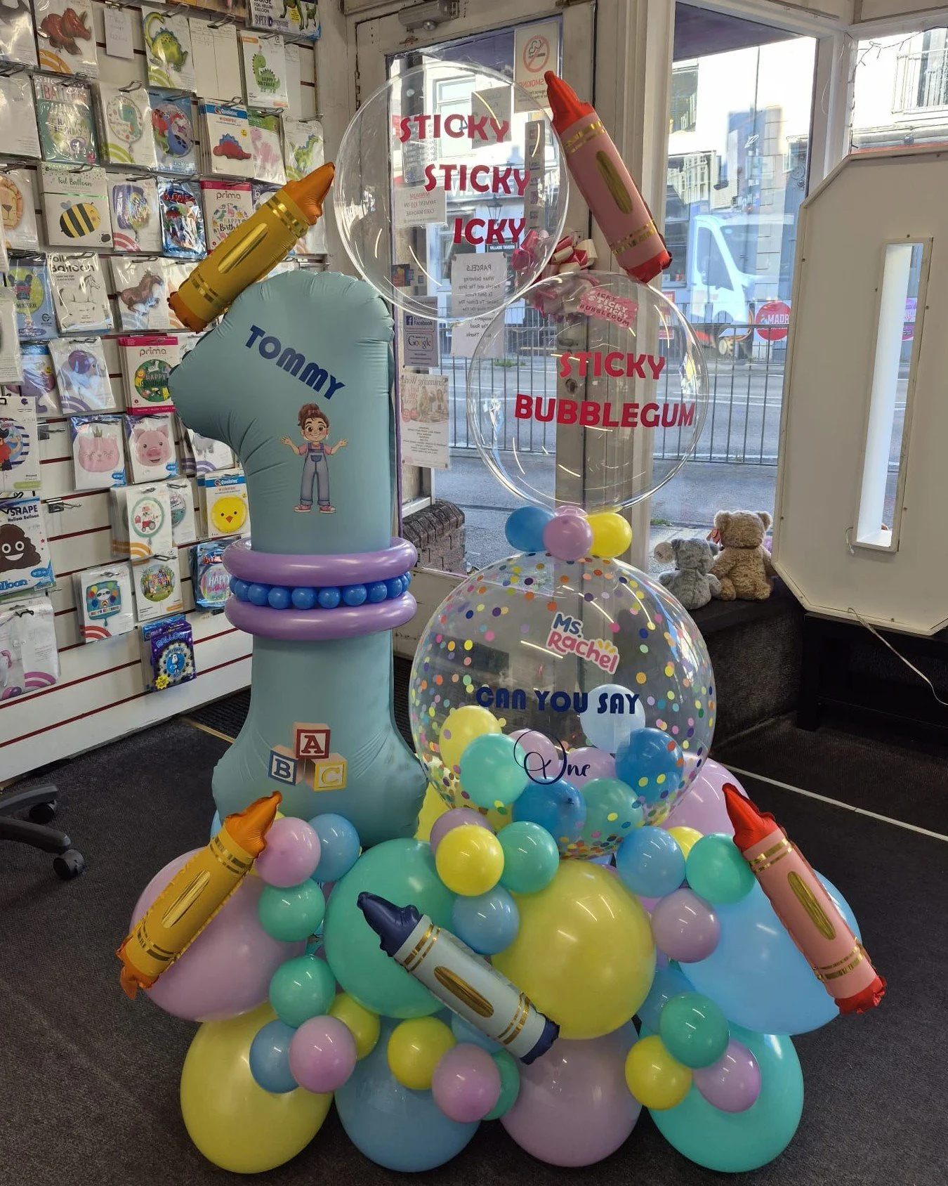 Colorful balloon display with toys and signs, including a large balloon with the words "CAN YOU SAY" and smaller balloons with children's names, in a store window.