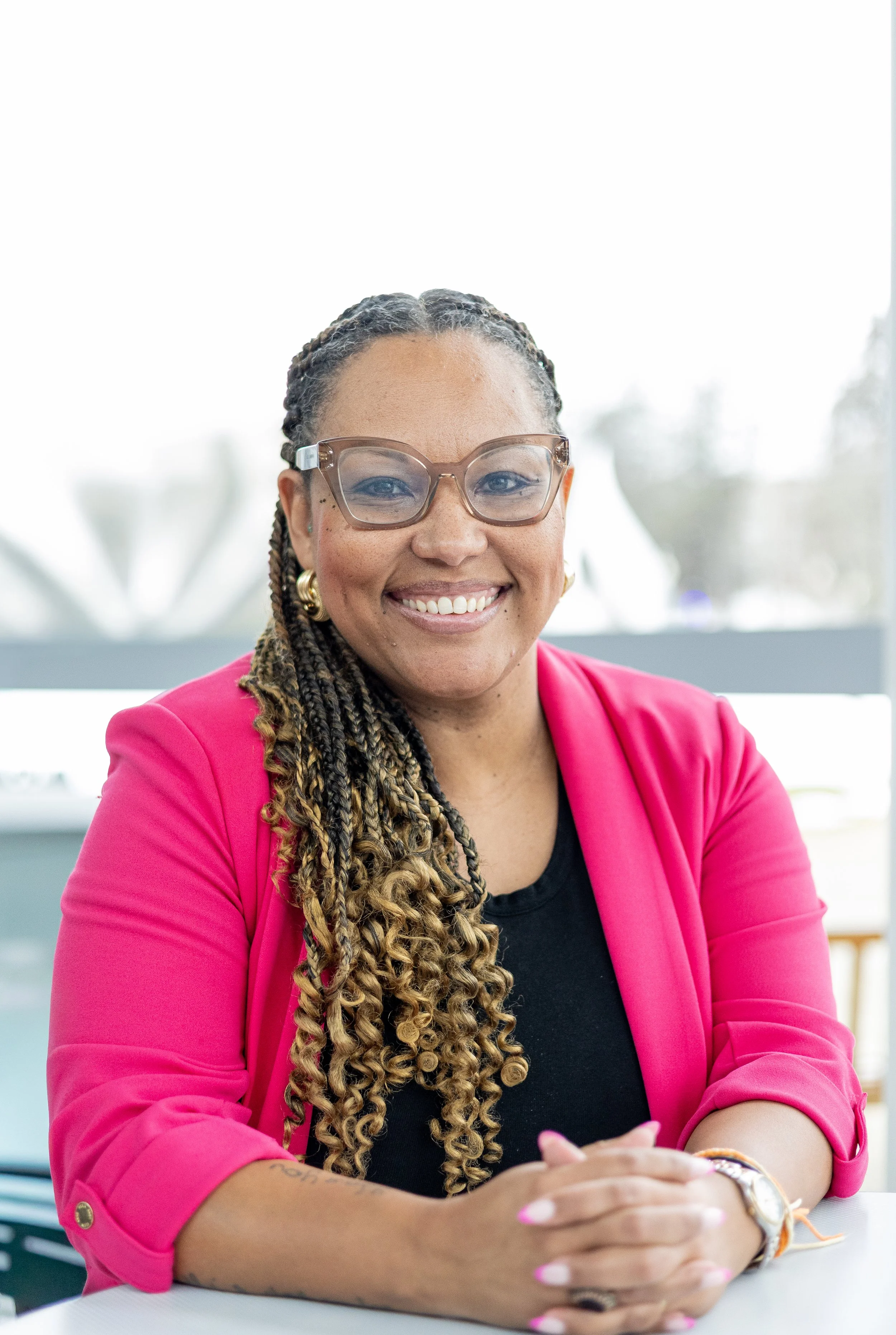 Black Cabo Verdean woman, lighter skin complexion, with a smile, long braids, brown glasses, gold earrings, black tshirt and pink blazer