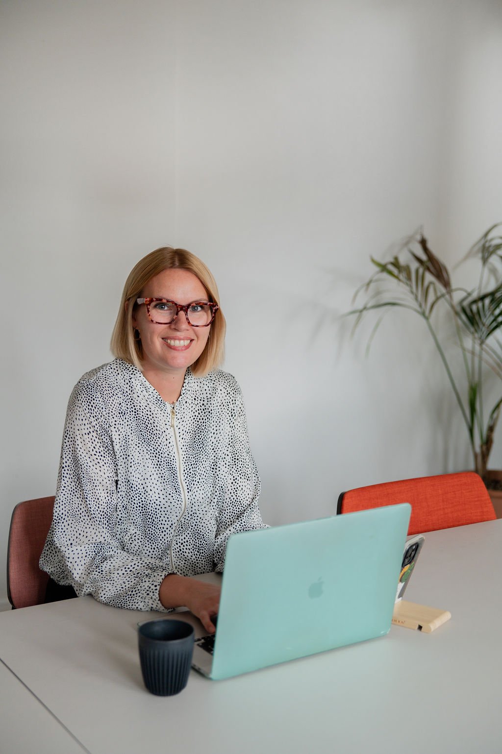 A woman with blonde hair and glasses sitting at a table with a laptop, a black mug, a yellow notebook, and a smartphone, smiling at the camera.