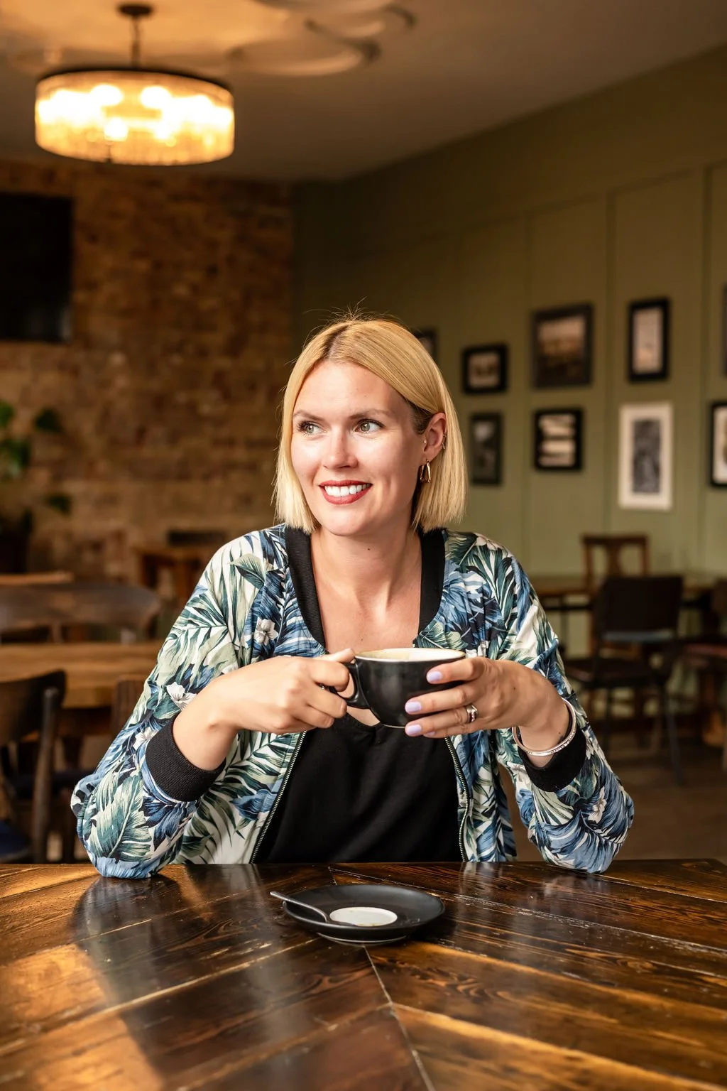 A woman with blonde hair holding a coffee cup, sitting at a wooden table in a cafe with artwork on the walls.