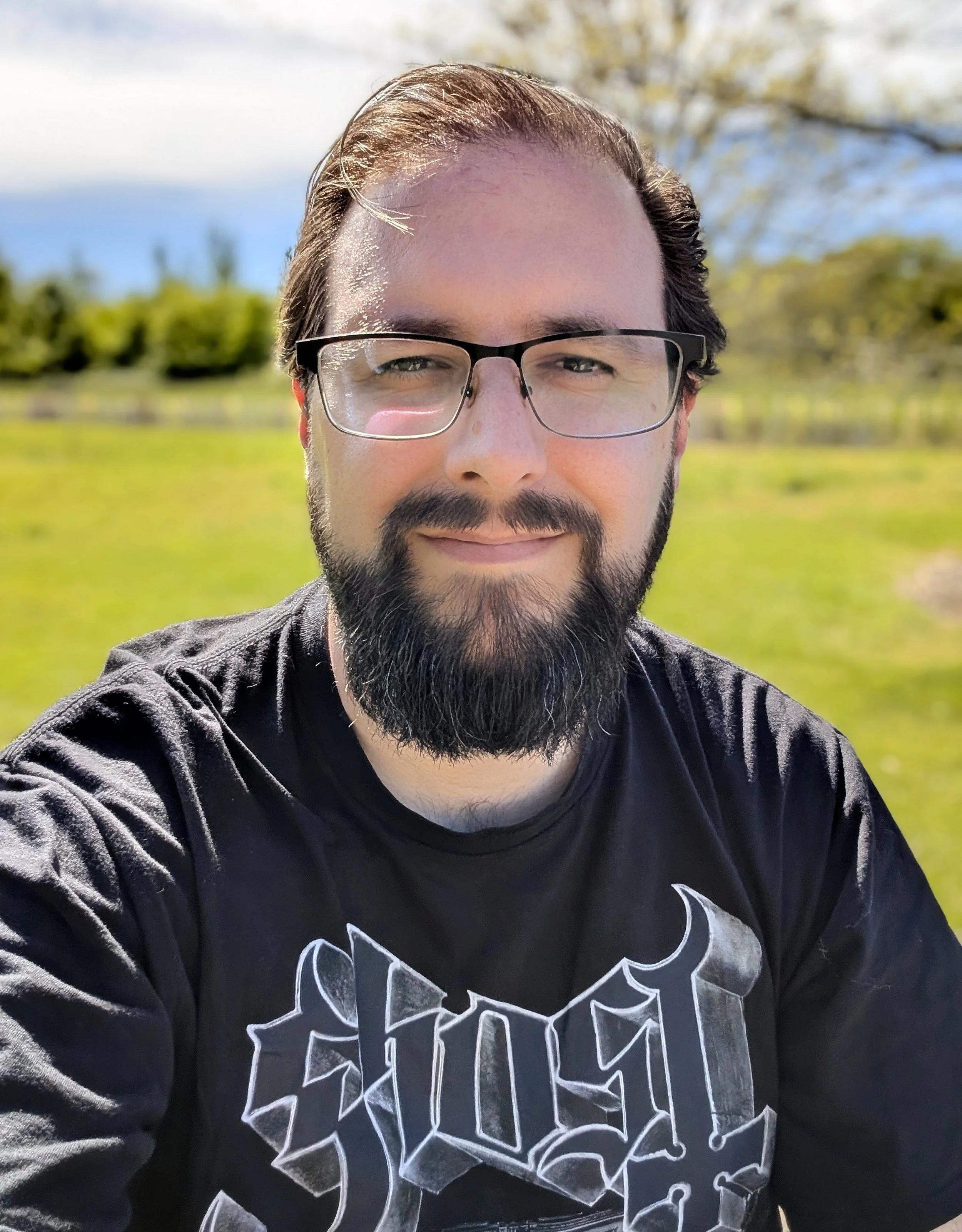 Bearded man with glasses wearing a black Ghost t-shirt outdoors.