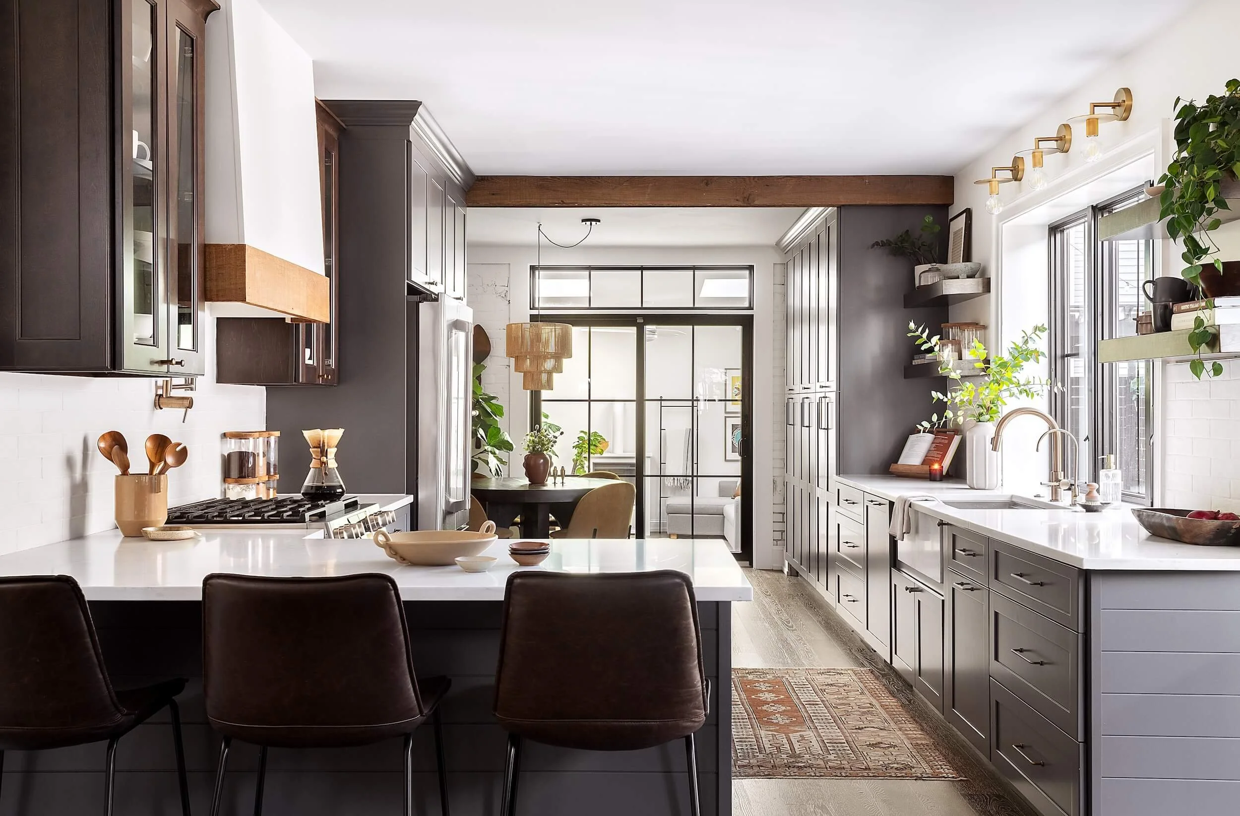 Sunlit kitchen with white marble counters, brass appliances, dark grey cabinets, and house plants
