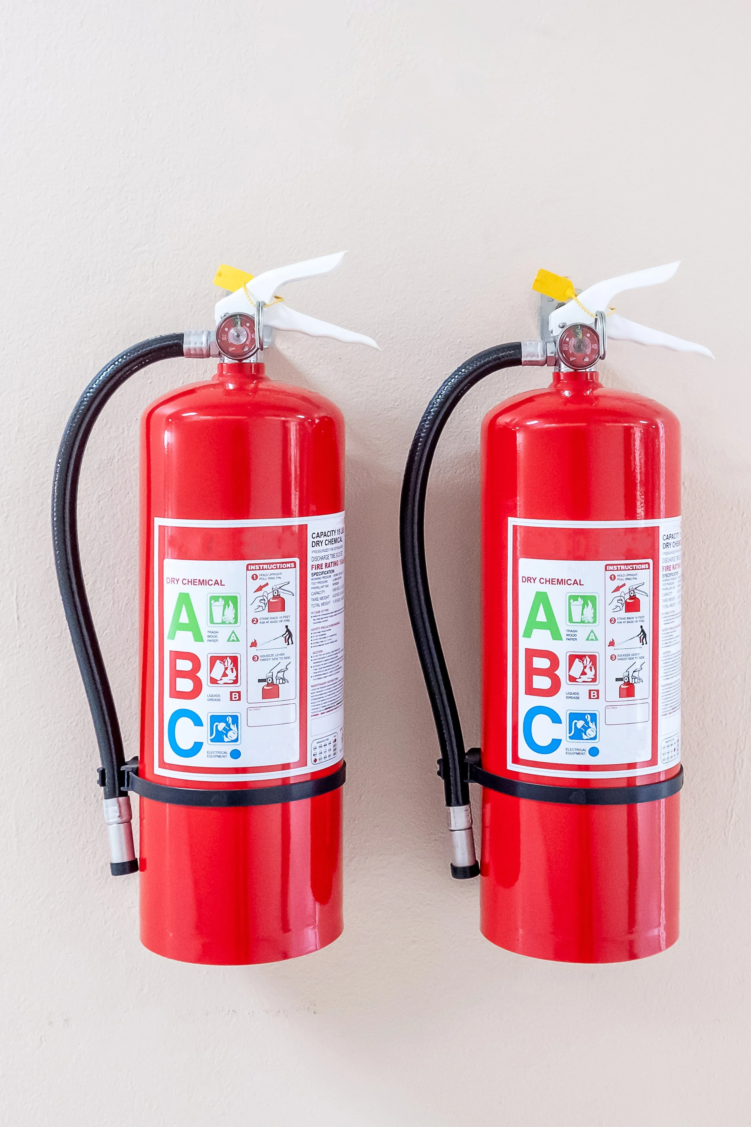 Two red fire extinguishers mounted on a beige wall, each with a white and yellow safety seal on the handle.