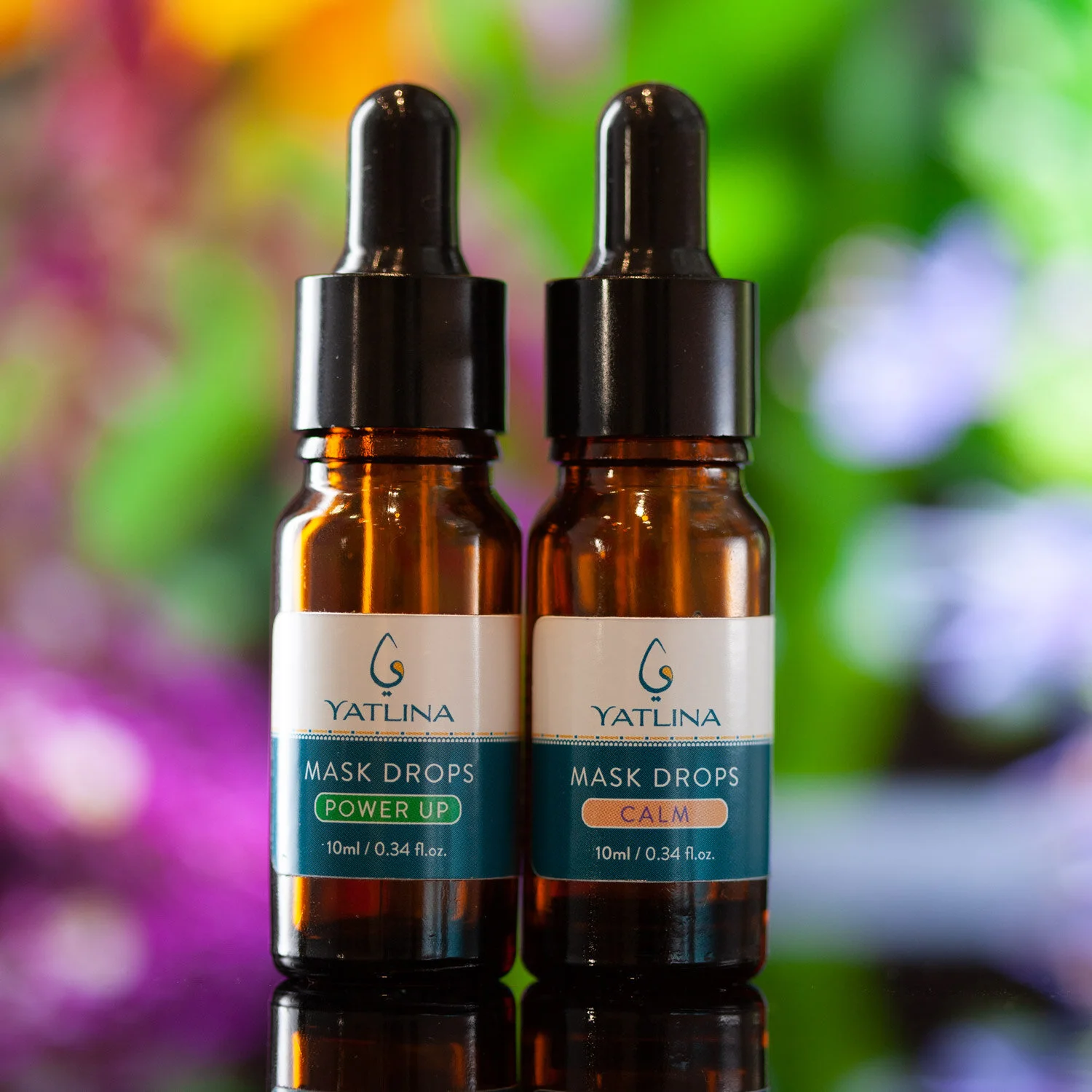 Two small amber glass bottles labeled 'Yatlina Mask Drops' with one labeled 'Power Up' and the other 'Calm,' featuring black dropper caps on a reflective surface with a blurred colorful floral background.